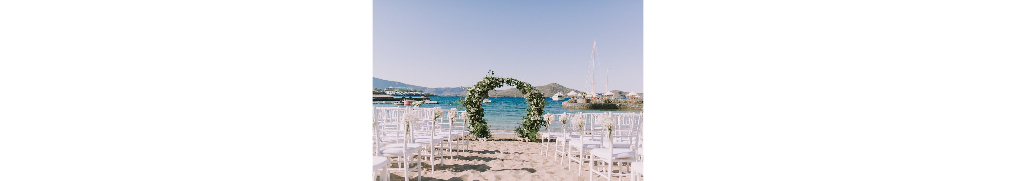 An elegant wedding ceremony setup is arranged on a sandy beach facing the ocean. White chairs line both sides of an aisle leading to a circular floral arch decorated with greenery and white flowers. Calm blue water, boats, and distant hills create a scenic coastal backdrop under a clear sky. The scene feels serene, romantic, and ready for guests to arrive.