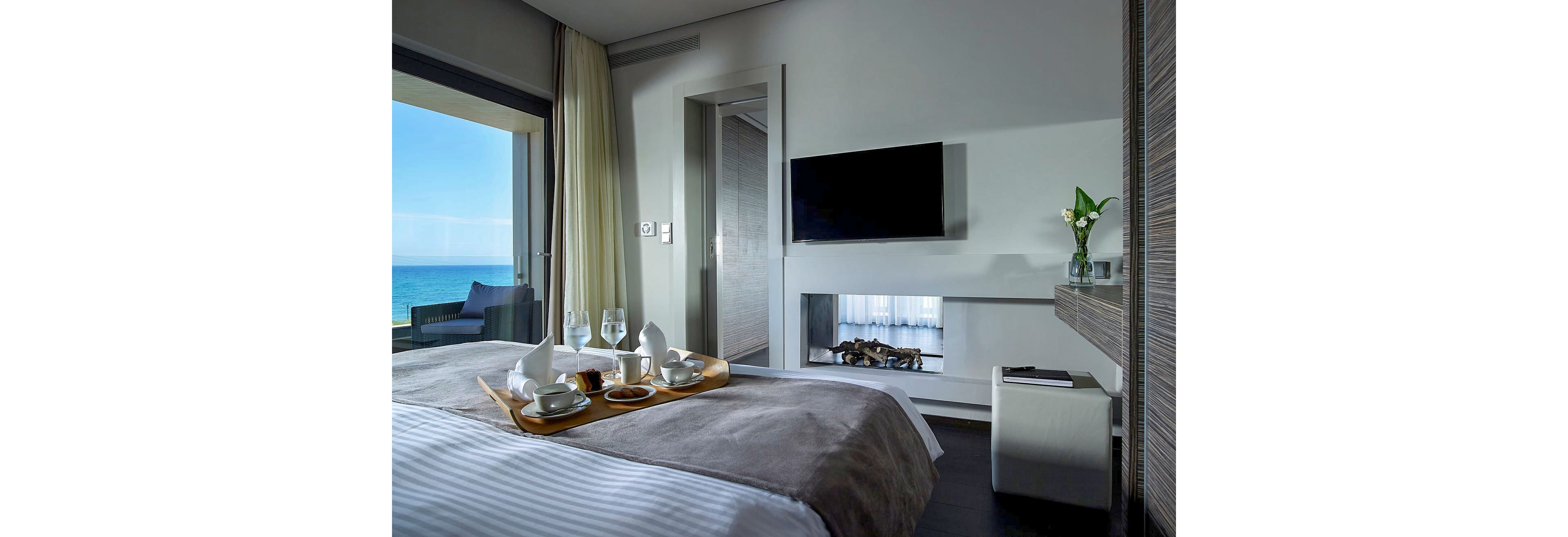 Contemporary hotel room features a neatly made bed with a breakfast tray holding cups, glasses, and plates. A wall-mounted flat-screen TV and built-in fireplace create a cozy, upscale atmosphere. Floor-to-ceiling curtains frame a balcony with seating and a clear view of the blue ocean outside. The overall style is minimalist and elegant, with cool tones and natural light enhancing the relaxing mood.
