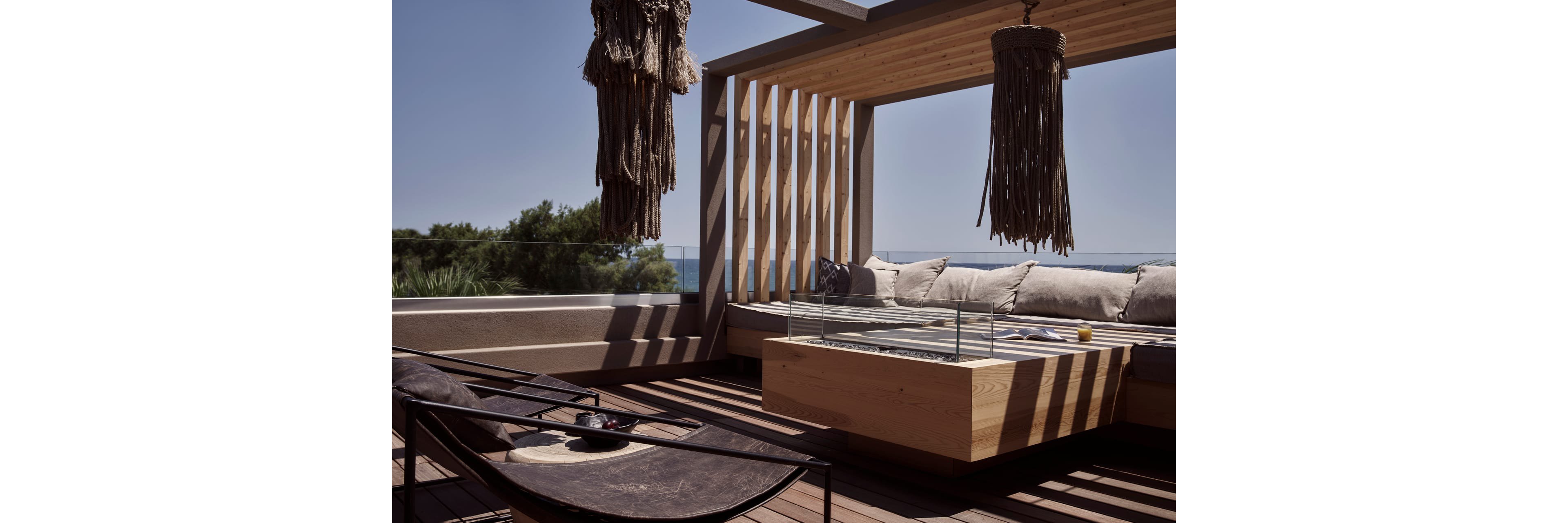 A contemporary outdoor lounge area features a built-in wooden sofa lined with neutral cushions under a slatted pergola. Sunlight filters through the wooden beams, casting shadows across the deck and low coffee table. Two large hanging woven light fixtures add texture and a relaxed, bohemian touch. In the background, trees and a distant view of the sea enhance the tranquil, resort-like atmosphere.