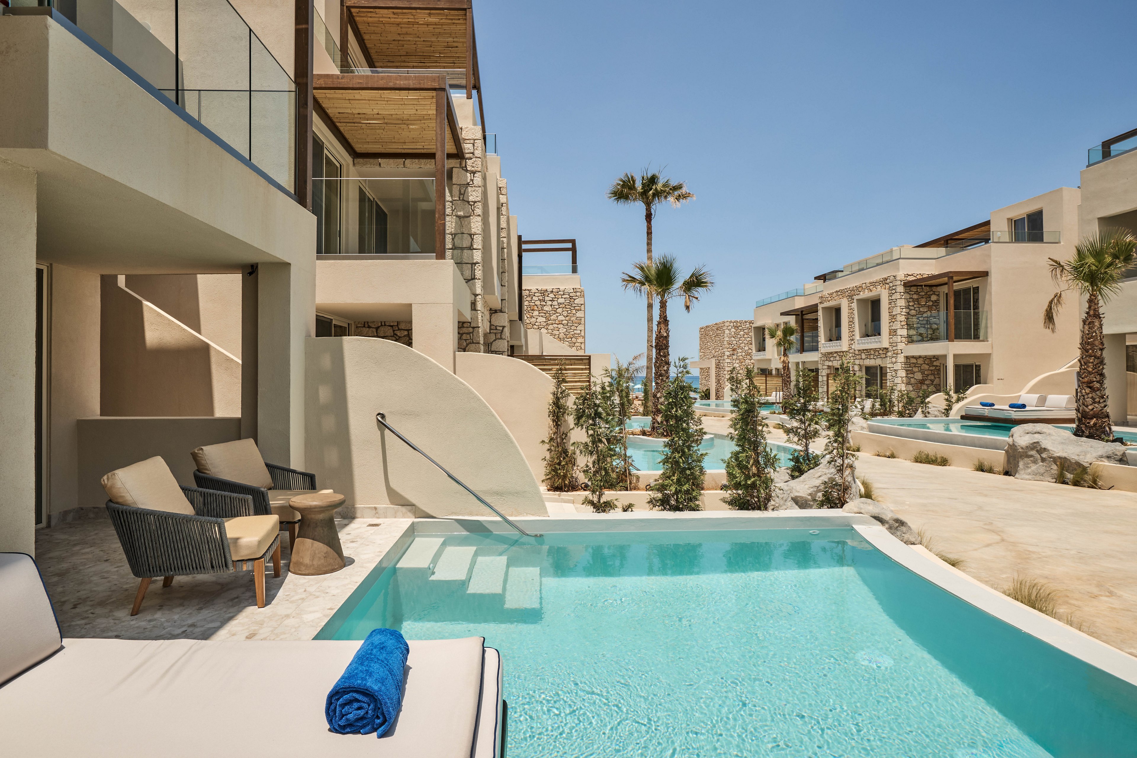 A serene poolside setting at a luxury resort featuring modern architectural design. The scene includes lounge chairs, a blue towel, and palm trees under a clear blue sky. Multiple pools and contemporary buildings create a tranquil atmosphere for relaxation.