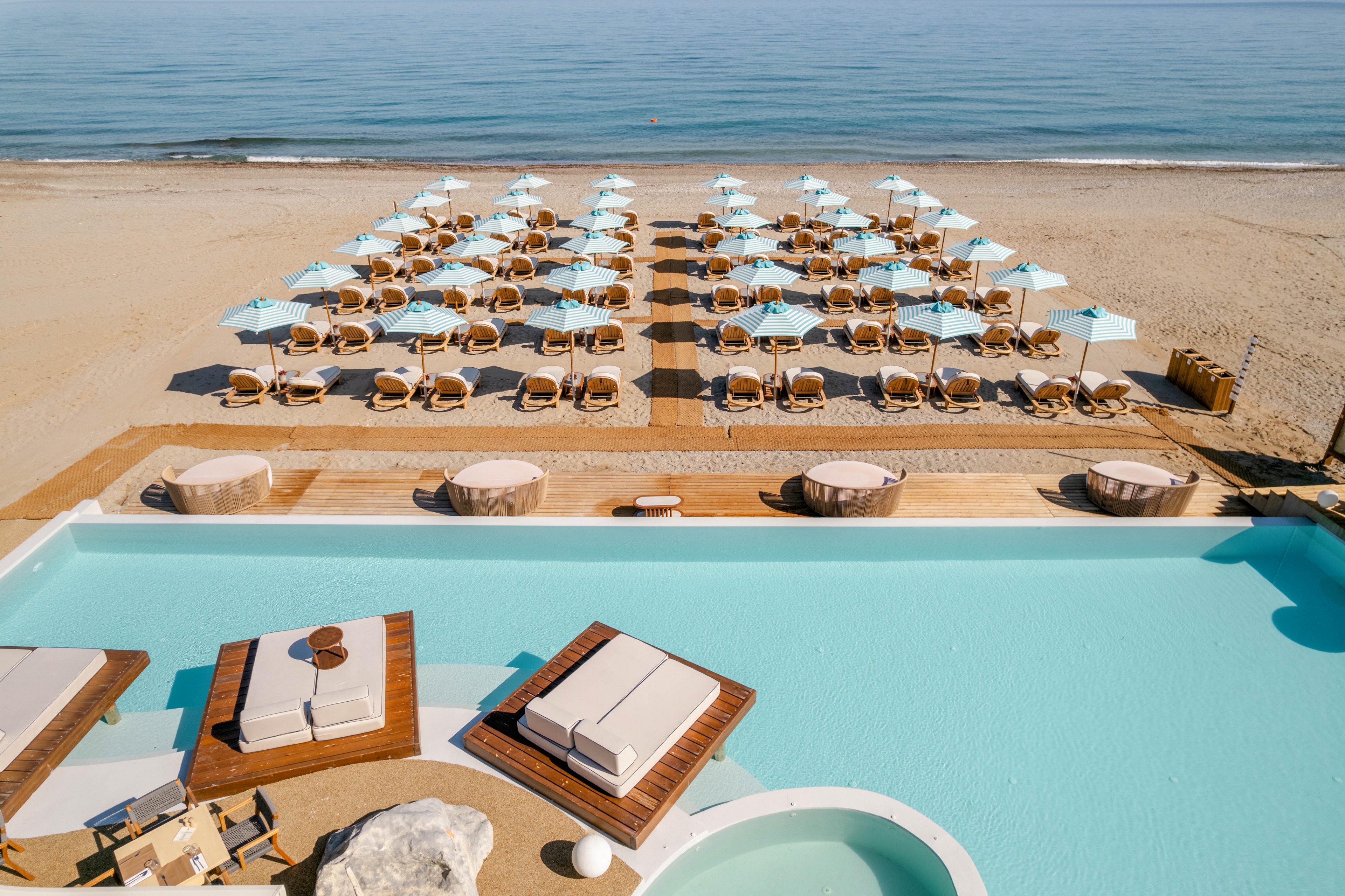 A luxurious beachfront resort features a serene pool in the foreground with cushioned daybeds on wooden platforms. Beyond the pool, rows of neatly arranged sun loungers and light blue umbrellas line the sandy beach. The calm sea stretches to the horizon under a clear sky, creating a tranquil coastal atmosphere. The scene emphasizes symmetry, relaxation, and modern seaside design.