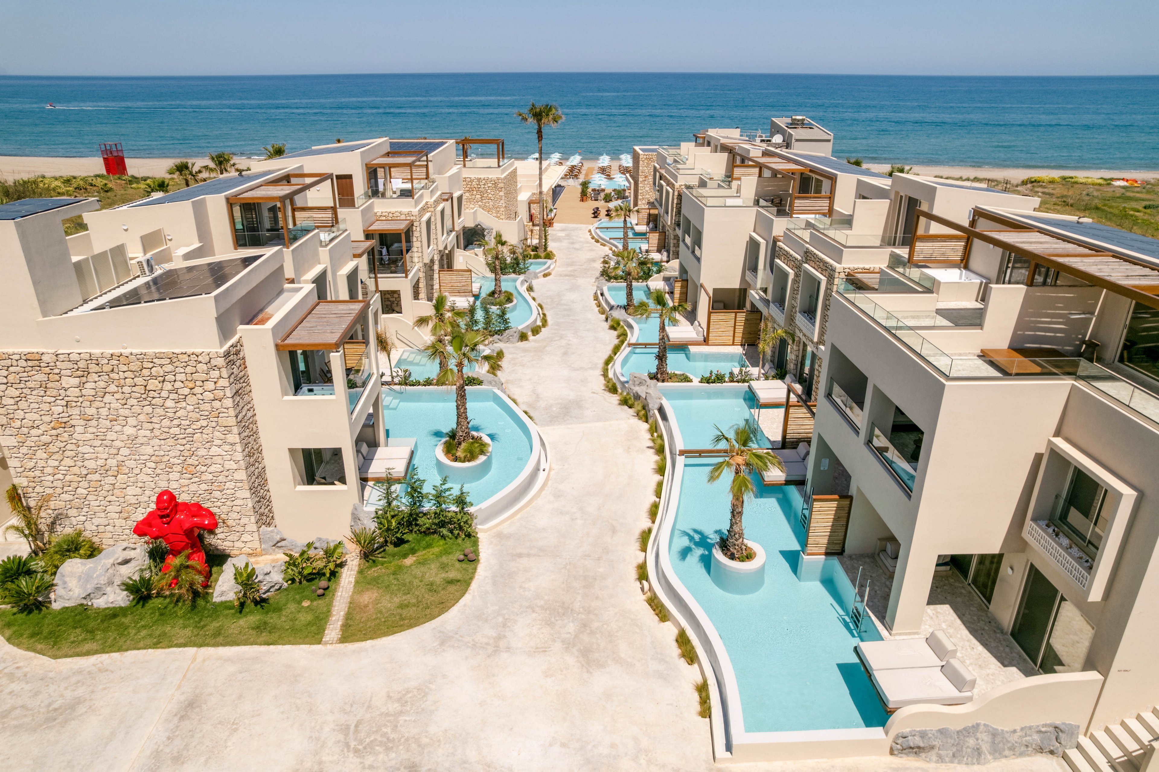 A contemporary beachfront resort features multiple beige low-rise buildings arranged around winding turquoise pools. The complex is situated directly beside a sandy beach and calm blue sea under a clear sky. Palm trees, sun loungers, and terraces create a relaxed vacation atmosphere. A large red sculpture stands near a stone wall, adding a bold artistic accent to the scene.