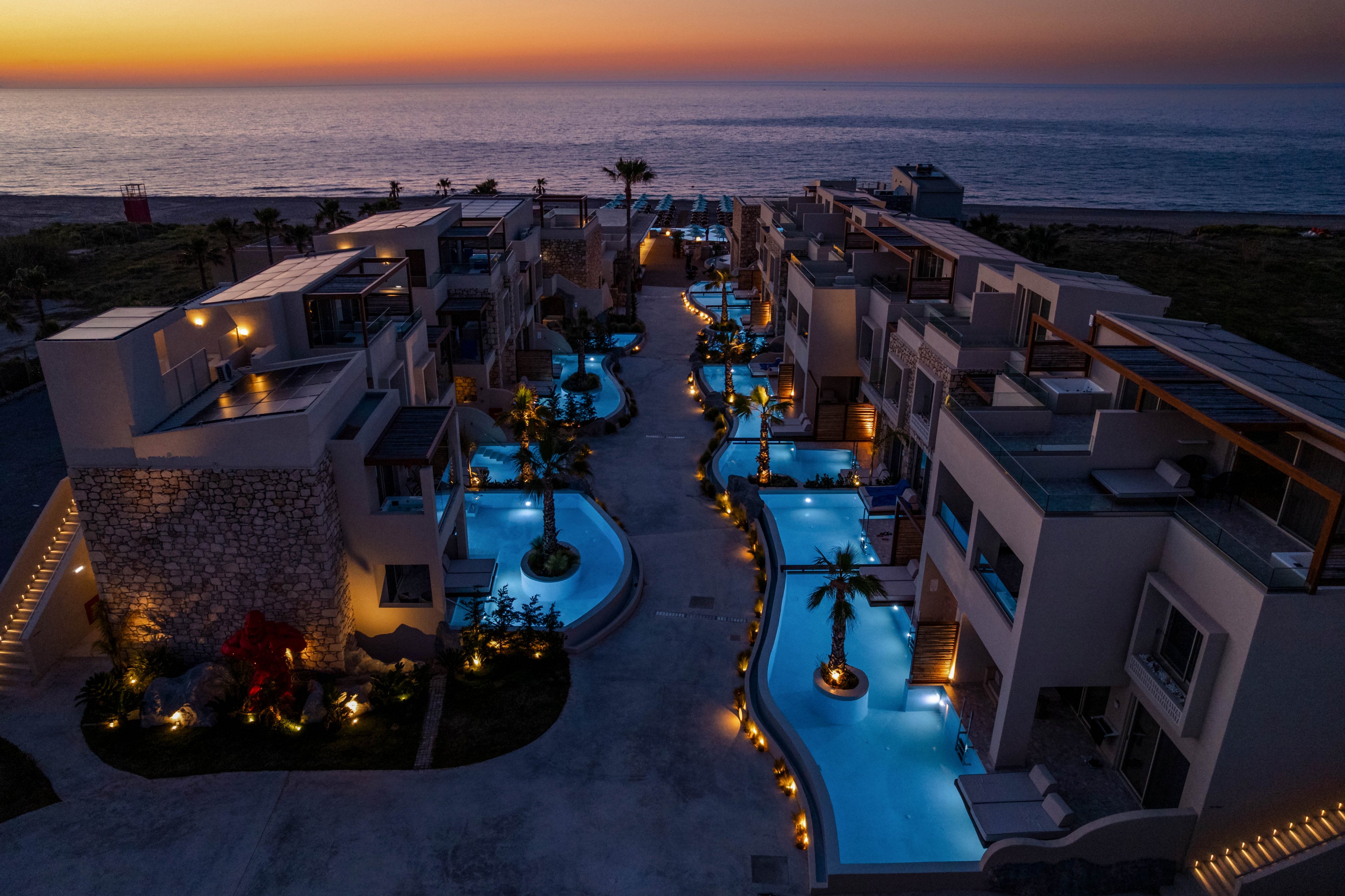 An aerial view captures a modern beachfront resort at dusk, with multiple illuminated swimming pools winding between contemporary buildings. The scene overlooks a calm ocean horizon under a colorful sunset sky. Palm trees, soft pathway lighting, and glowing blue water create a serene, upscale atmosphere. The overall mood is tranquil and inviting, suggesting a premium vacation destination.