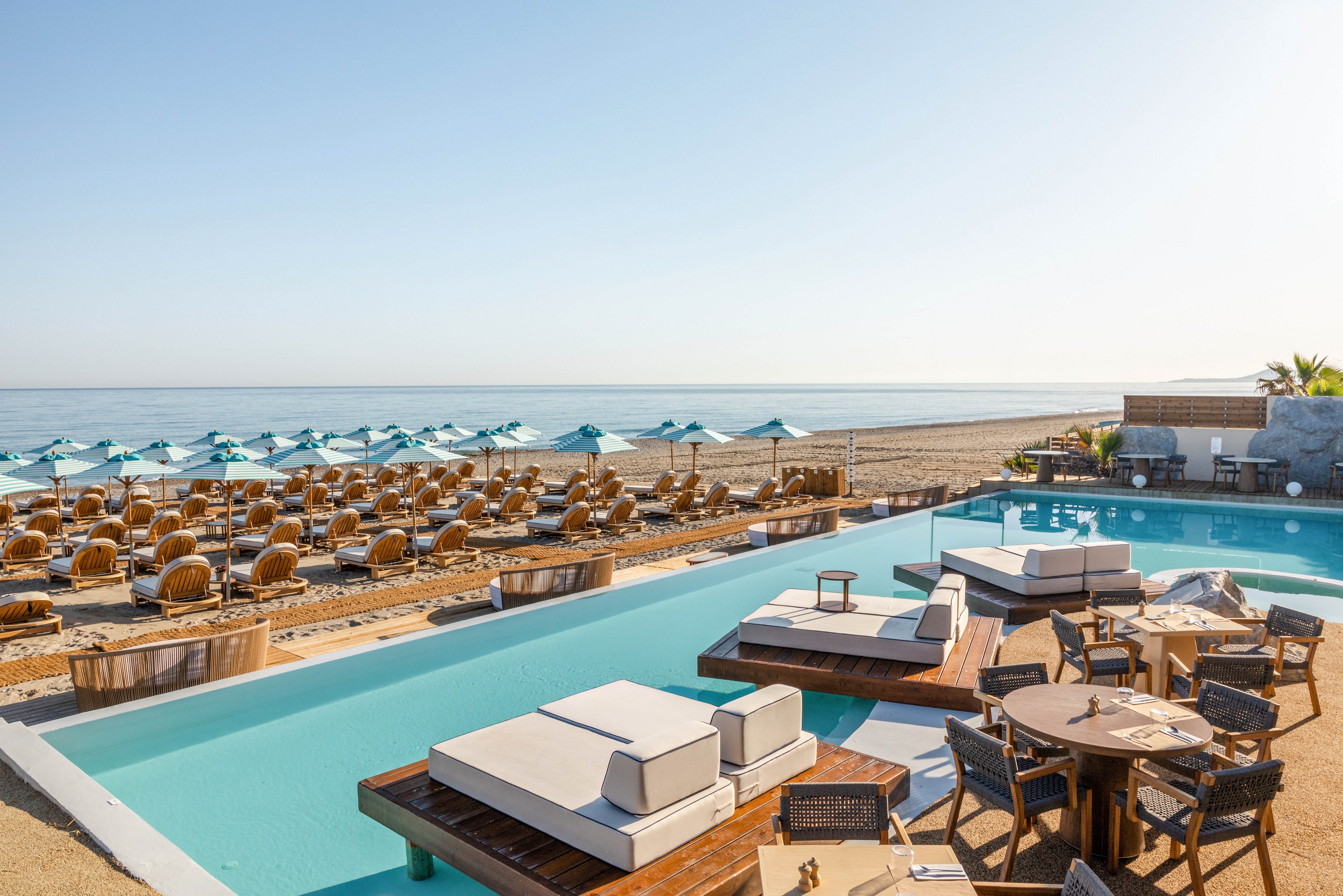 A modern beachfront resort features a sleek swimming pool with cushioned lounge platforms and wooden sunbeds. Rows of beach chairs and teal umbrellas line the sandy shore facing the calm sea. Outdoor dining tables and chairs sit beside the pool, creating a relaxed vacation atmosphere. The clear sky and bright sunlight emphasize the tranquil, minimalist coastal setting.