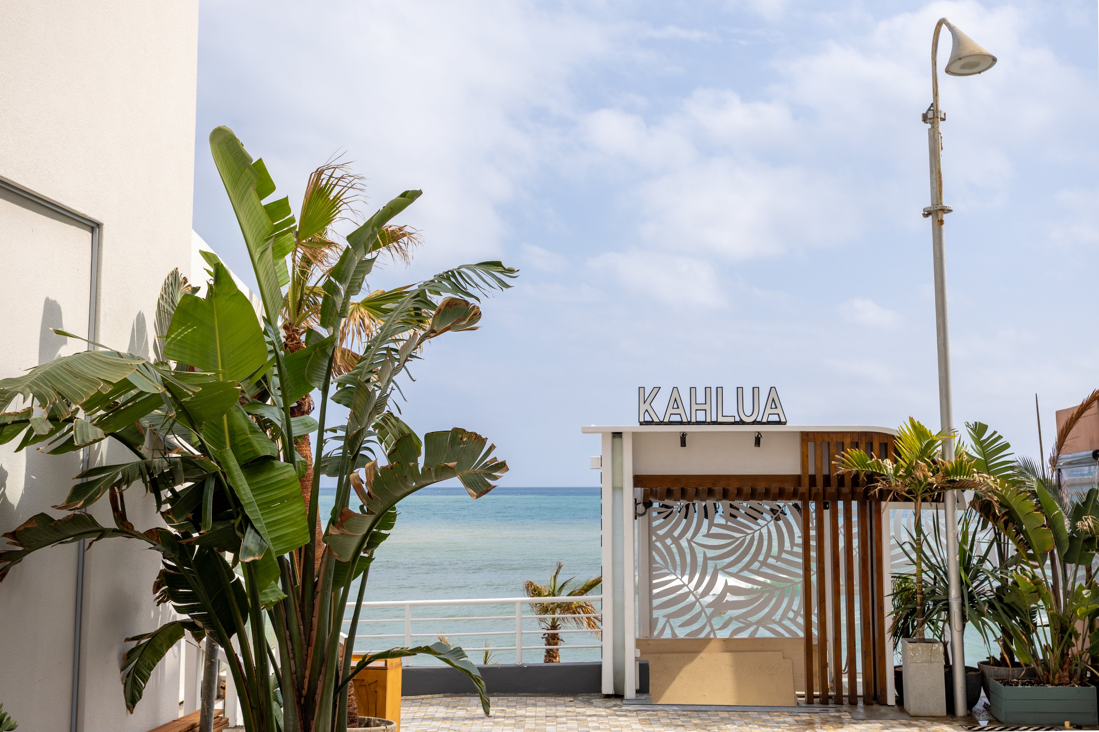 A small beachfront bar named KAHLUA stands facing a calm blue ocean. Tropical plants and palm trees frame the entrance, creating a relaxed coastal atmosphere. The bar structure features light wood, white panels, and a decorative patterned screen. A clear sky with scattered clouds and a tall street lamp complete the seaside setting.