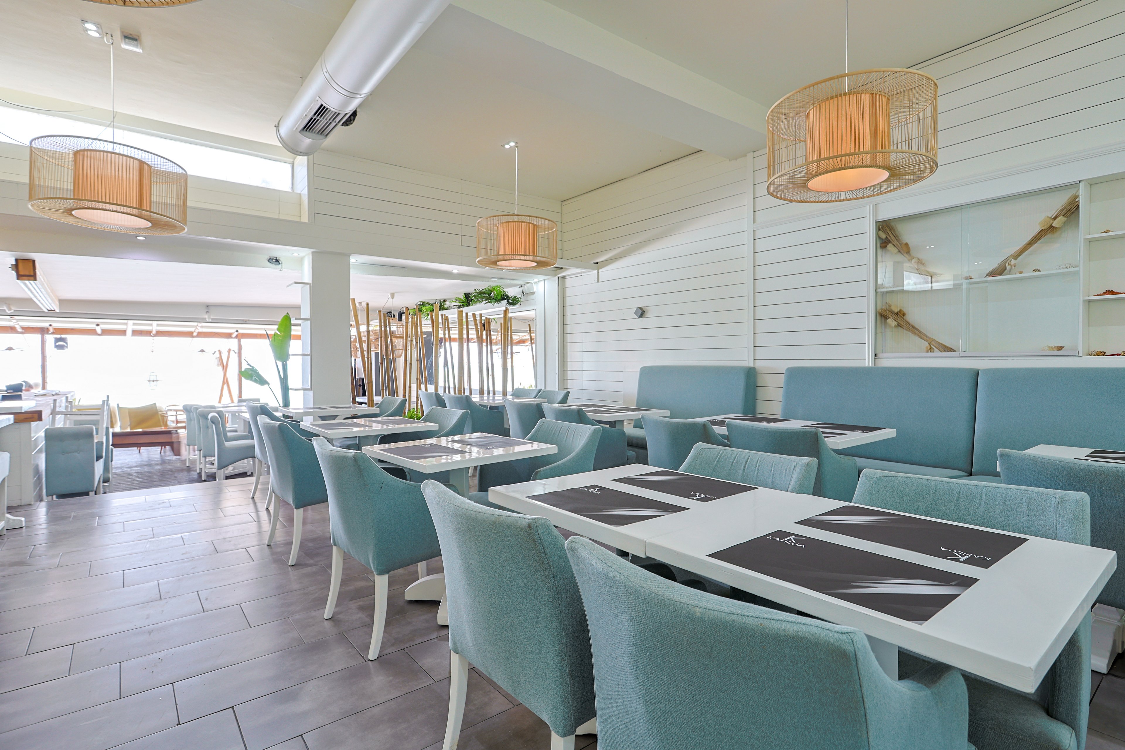 A modern restaurant interior features rows of teal upholstered chairs and banquettes around white tables. Large windows and glass doors let in abundant natural light, creating an airy, open atmosphere. Pendant lights with woven shades hang from the ceiling, complementing the light wood and white wall paneling. The space appears clean, minimalist, and ready to welcome guests in a relaxed coastal setting.