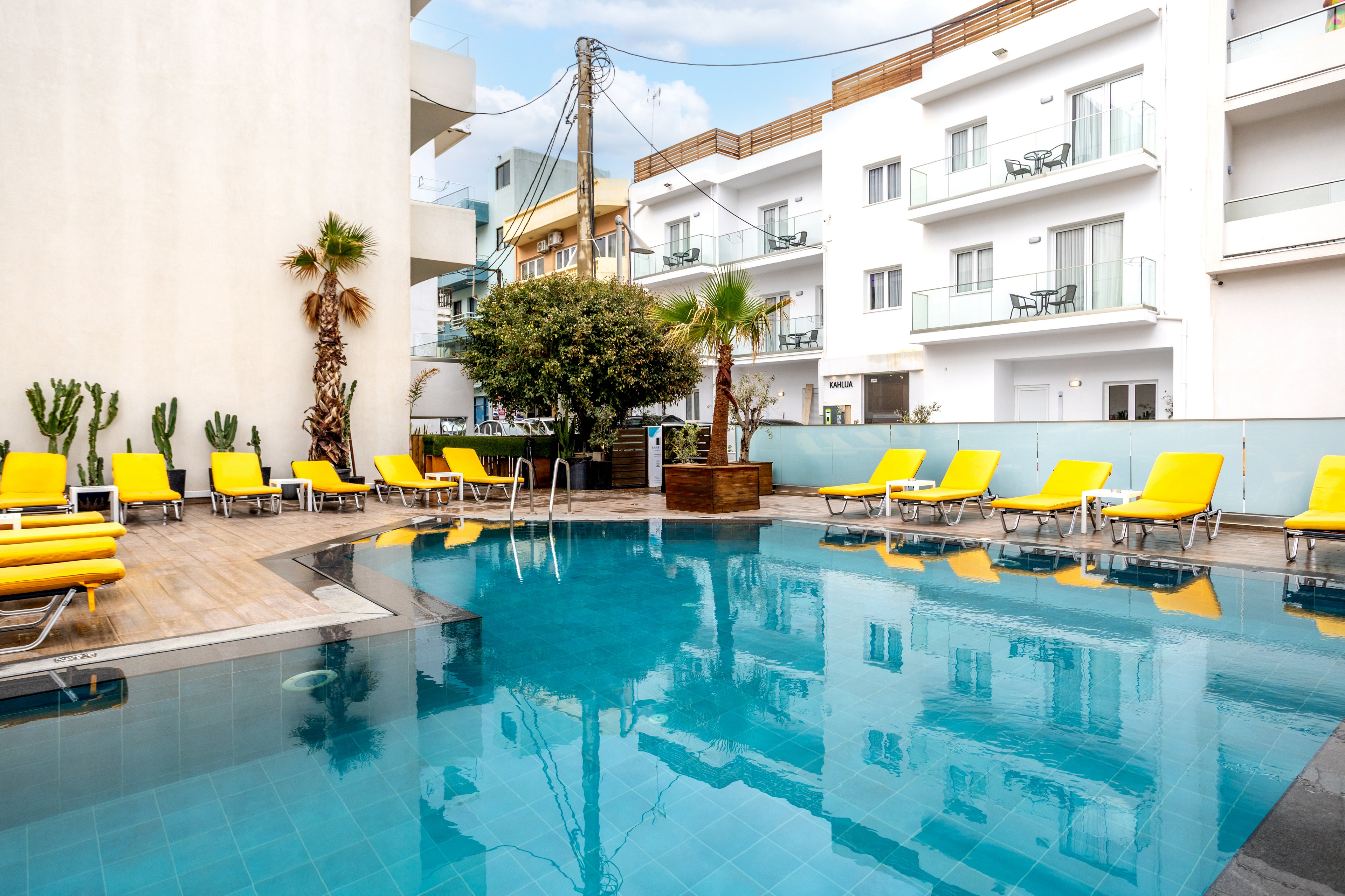 A clear blue swimming pool sits in the courtyard of a modern hotel or apartment complex. Bright yellow sun loungers line the poolside on wooden decking, creating a vibrant contrast with the blue water. Palm trees and potted plants add a touch of greenery to the urban setting. White residential-style buildings with balconies surround the pool area under a partly cloudy sky.