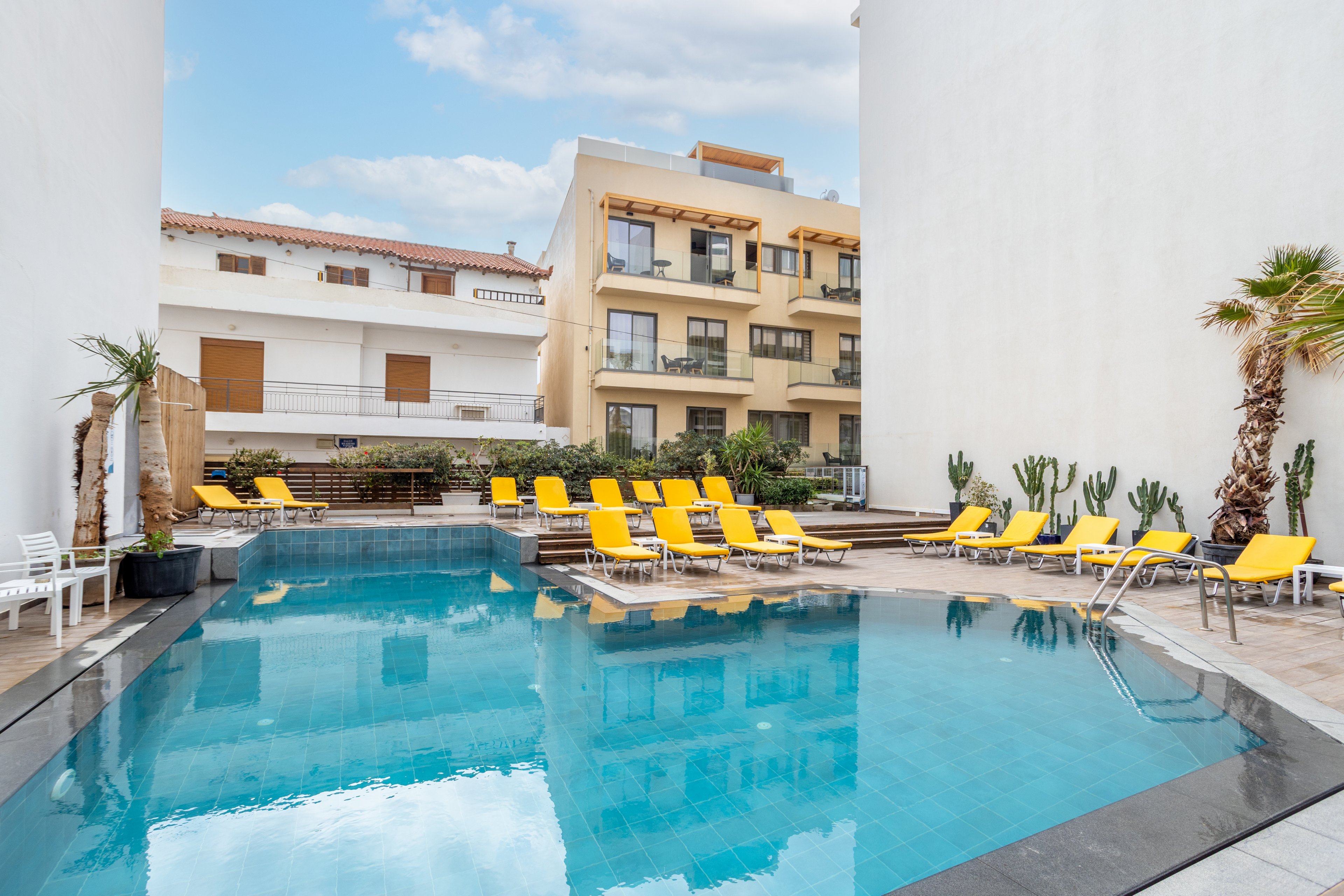 A clear blue swimming pool sits in the courtyard between modern white buildings. Bright yellow lounge chairs line the poolside, creating a vibrant contrast against the neutral architecture. Potted cacti and a few palm trees add a touch of greenery and a warm-climate feel. The calm water and empty seating suggest a quiet, relaxing atmosphere in an urban hotel setting.