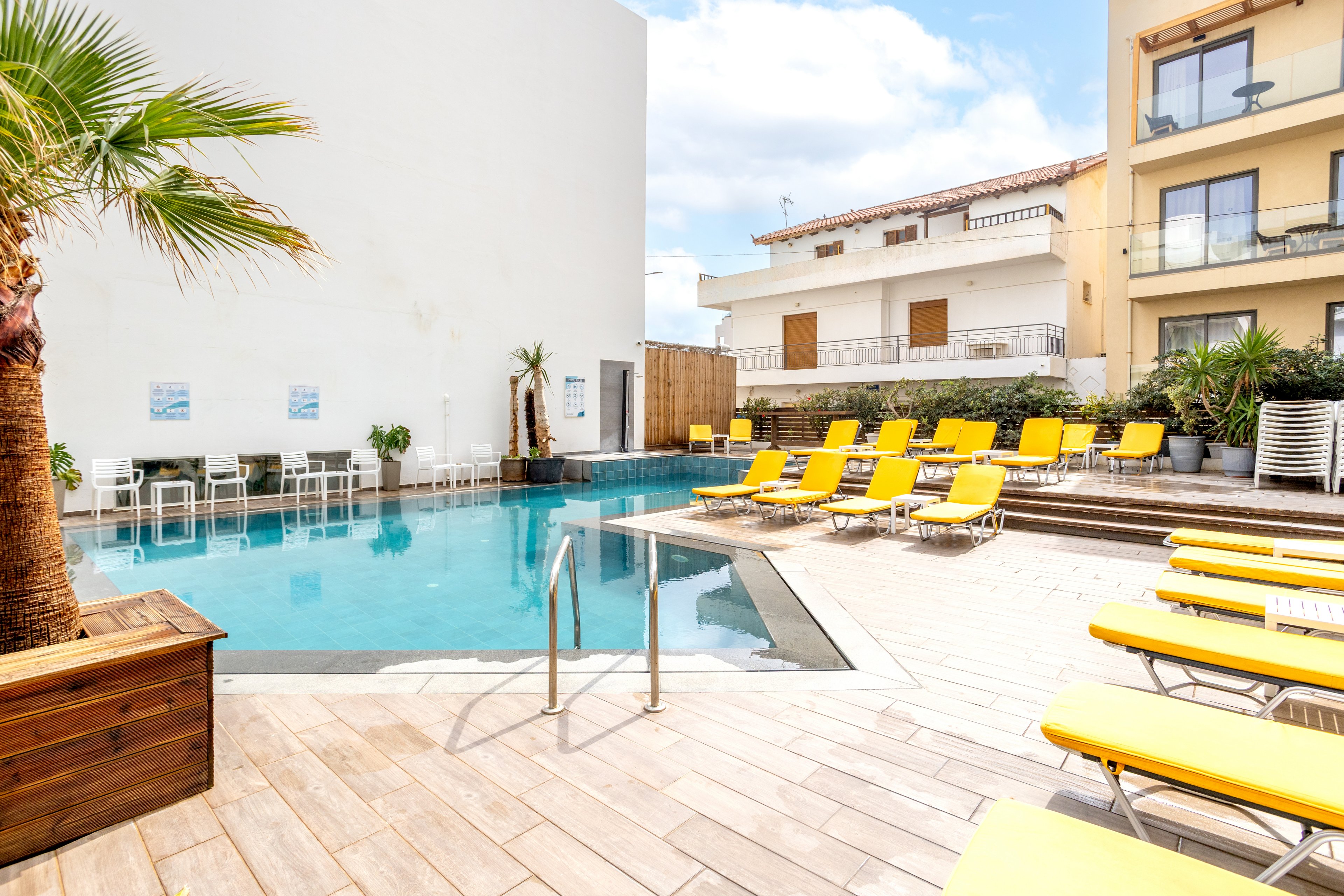 An outdoor swimming pool is surrounded by bright yellow sun loungers in a modern hotel courtyard. The clear blue water reflects the sky and nearby buildings, creating a calm and inviting atmosphere. Palm trees and potted plants add a touch of greenery to the wooden deck area. Residential-style balconies and white walls frame the pool, suggesting a relaxed vacation setting.