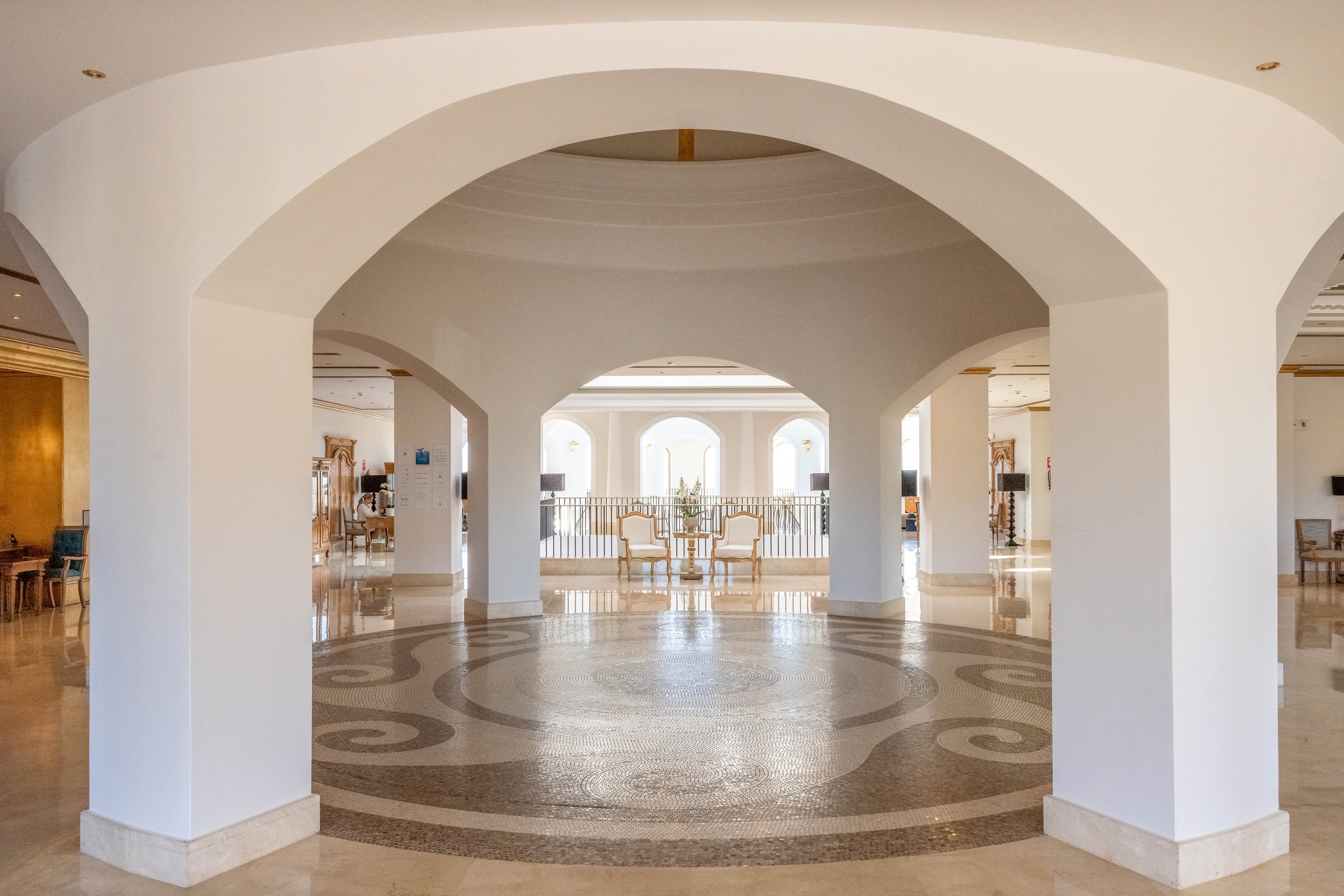 A spacious hotel lobby features large white arches framing an open circular foyer. Natural light floods in through tall arched windows at the far end, illuminating the polished stone floor with decorative patterns. Elegant seating and tables are arranged near the balcony railing, suggesting a relaxed lounge or reception area. The overall scene feels airy, refined, and welcoming.