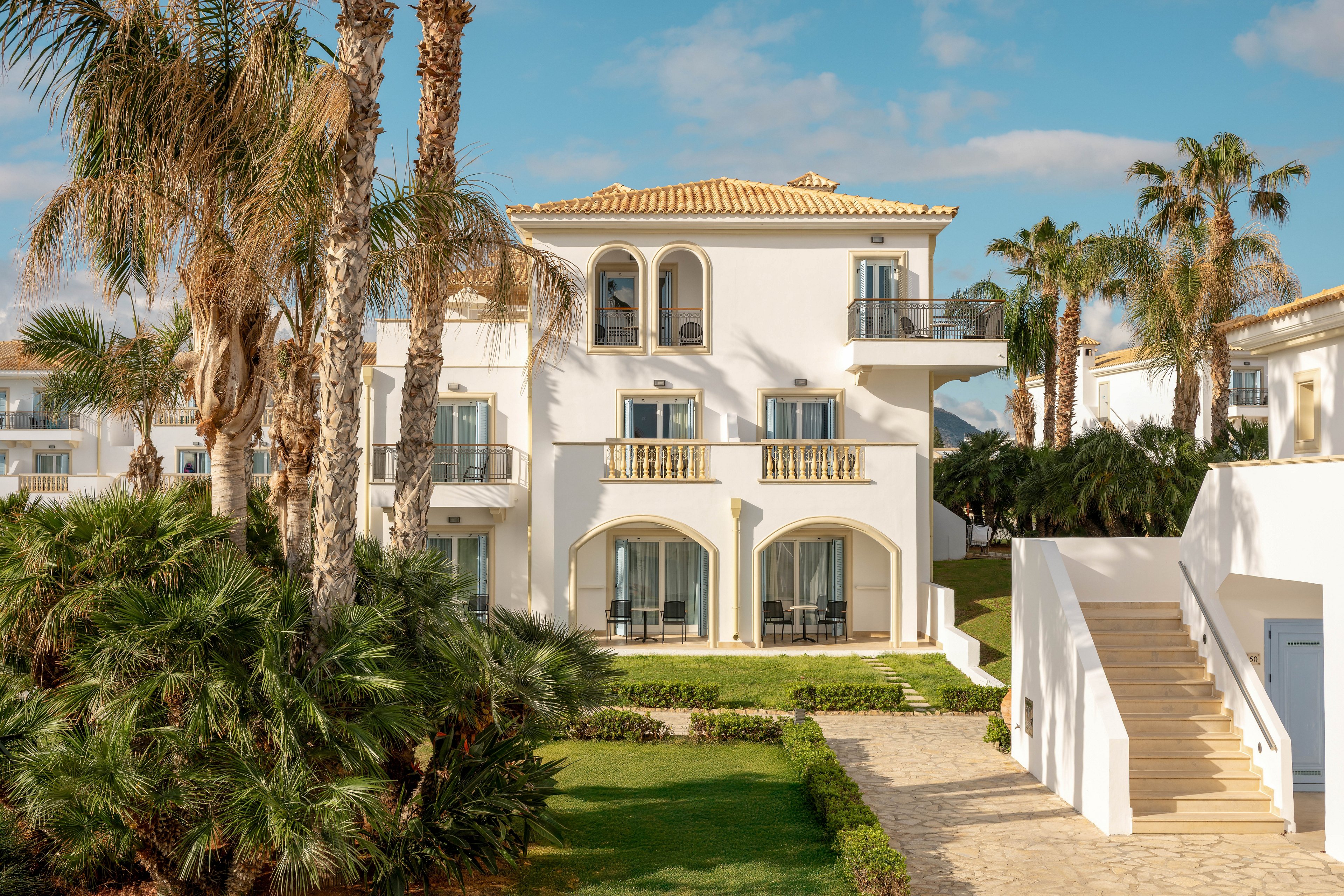 A white Mediterranean-style resort building stands surrounded by lush palm trees and manicured gardens. The structure features arched windows, balconies with railings, and a tiled roof under a bright blue sky. A stone pathway and exterior staircase lead to the entrances, suggesting guest rooms or vacation apartments. Warm sunlight enhances the cream and beige tones of the architecture, creating a relaxed holiday atmosphere.
