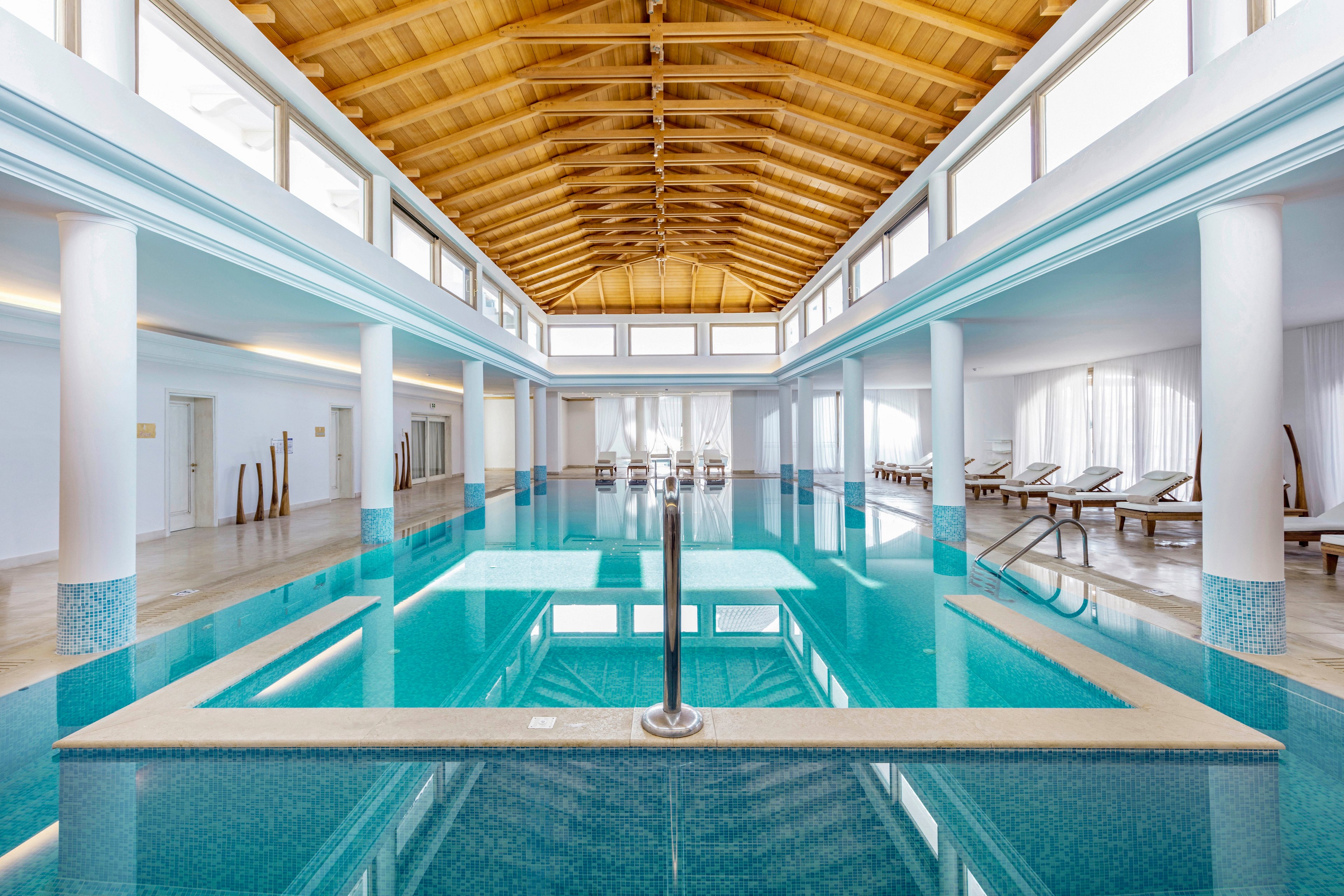 A serene indoor swimming pool stretches beneath a high wooden ceiling supported by white columns. The clear turquoise water reflects the architecture, creating a calm and symmetrical scene. Sun loungers line the sides of the pool near large windows and sheer curtains, suggesting a relaxing spa or resort environment. Soft natural light and clean white walls enhance the tranquil, minimalist atmosphere.
