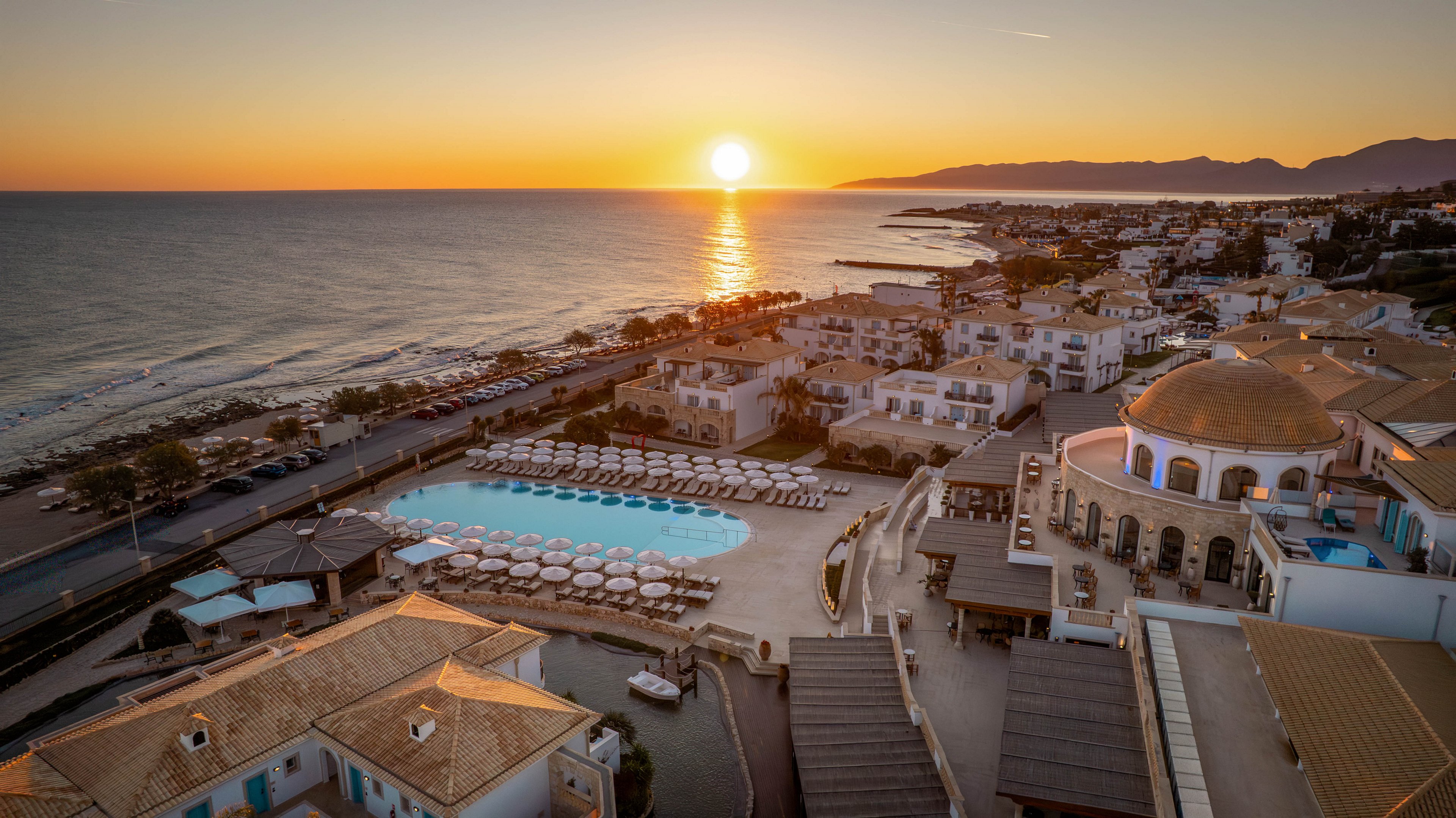 An aerial view captures a seaside resort at sunset, with the sun low over the horizon. A large swimming pool surrounded by rows of sun loungers and umbrellas forms the central focus of the complex. The resort buildings feature light-colored roofs and a prominent domed structure near the pool area. The coastline, calm sea, and distant mountains create a scenic backdrop, while the warm golden light enhances the tranquil atmosphere.