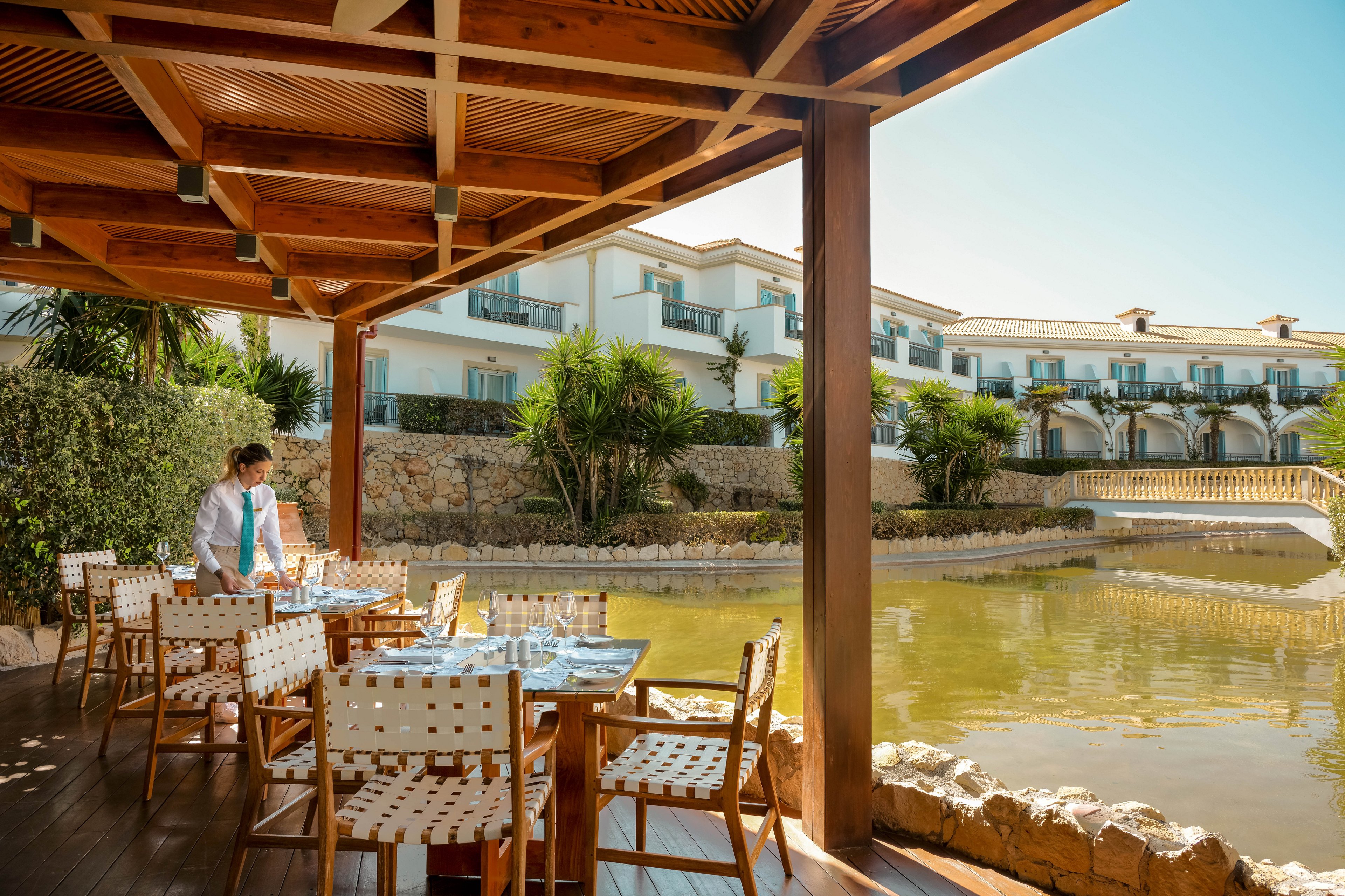 A waiter prepares a table at an outdoor restaurant terrace beside a calm artificial lake. The covered wooden deck features woven chairs and neatly set tables with glassware. In the background, a modern white resort hotel with balconies overlooks the water and lush greenery. Warm sunlight and clear skies create a relaxed vacation atmosphere.
