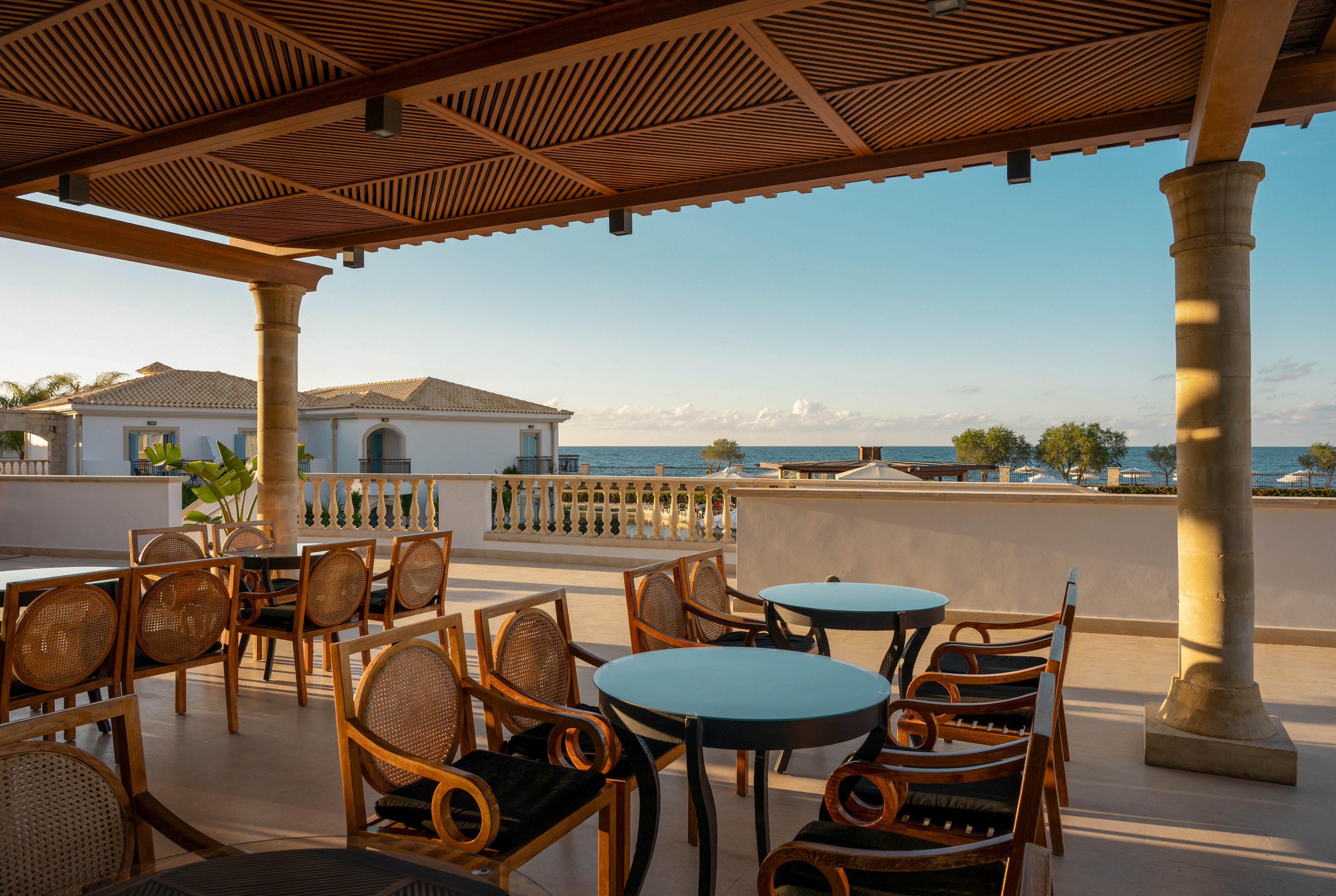 An open-air terrace cafe overlooks a calm seaside resort. Wooden chairs and round tables are arranged under a shaded pergola with warm afternoon light. Elegant columns and balustrades frame the view of white villas and the distant ocean. The scene feels tranquil and inviting, ideal for relaxing or dining outdoors.