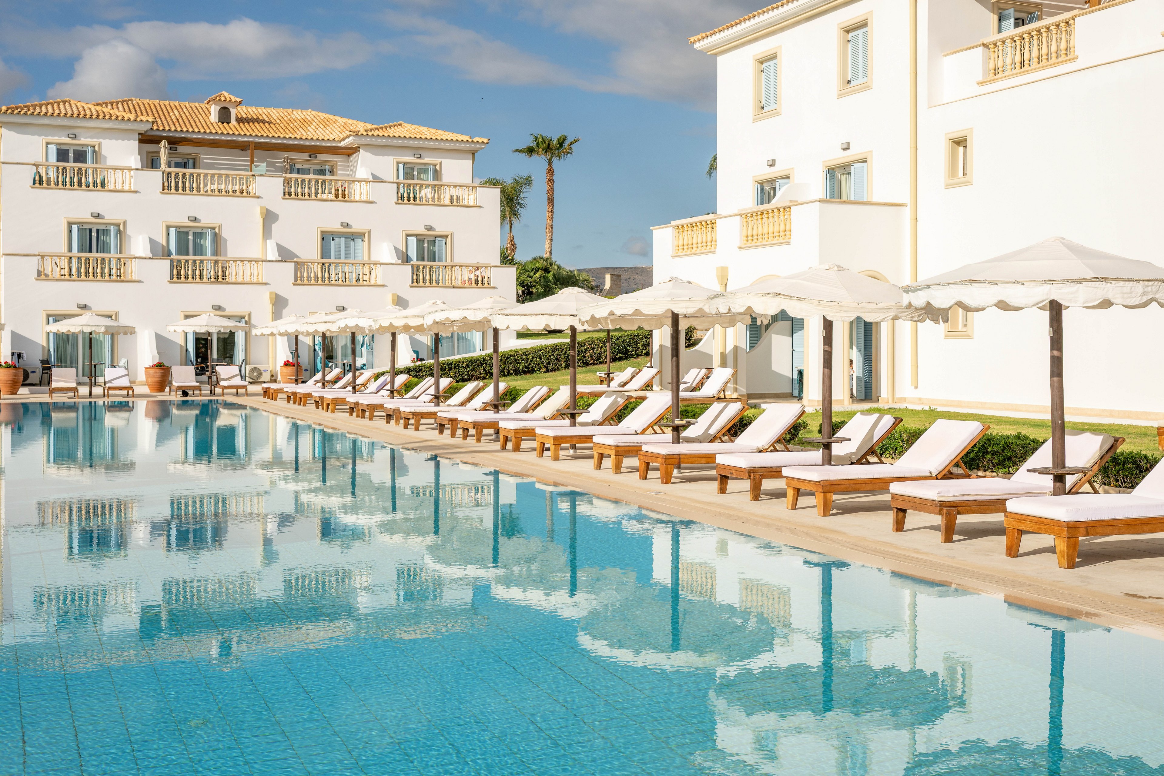 A luxurious resort pool is lined with white cushioned loungers and large sun umbrellas. The clear blue water reflects the bright white hotel building and balconies in the background. Palm trees and a blue sky with scattered clouds enhance the tropical vacation atmosphere. The scene feels calm, sunny, and inviting, suggesting a relaxing holiday destination.