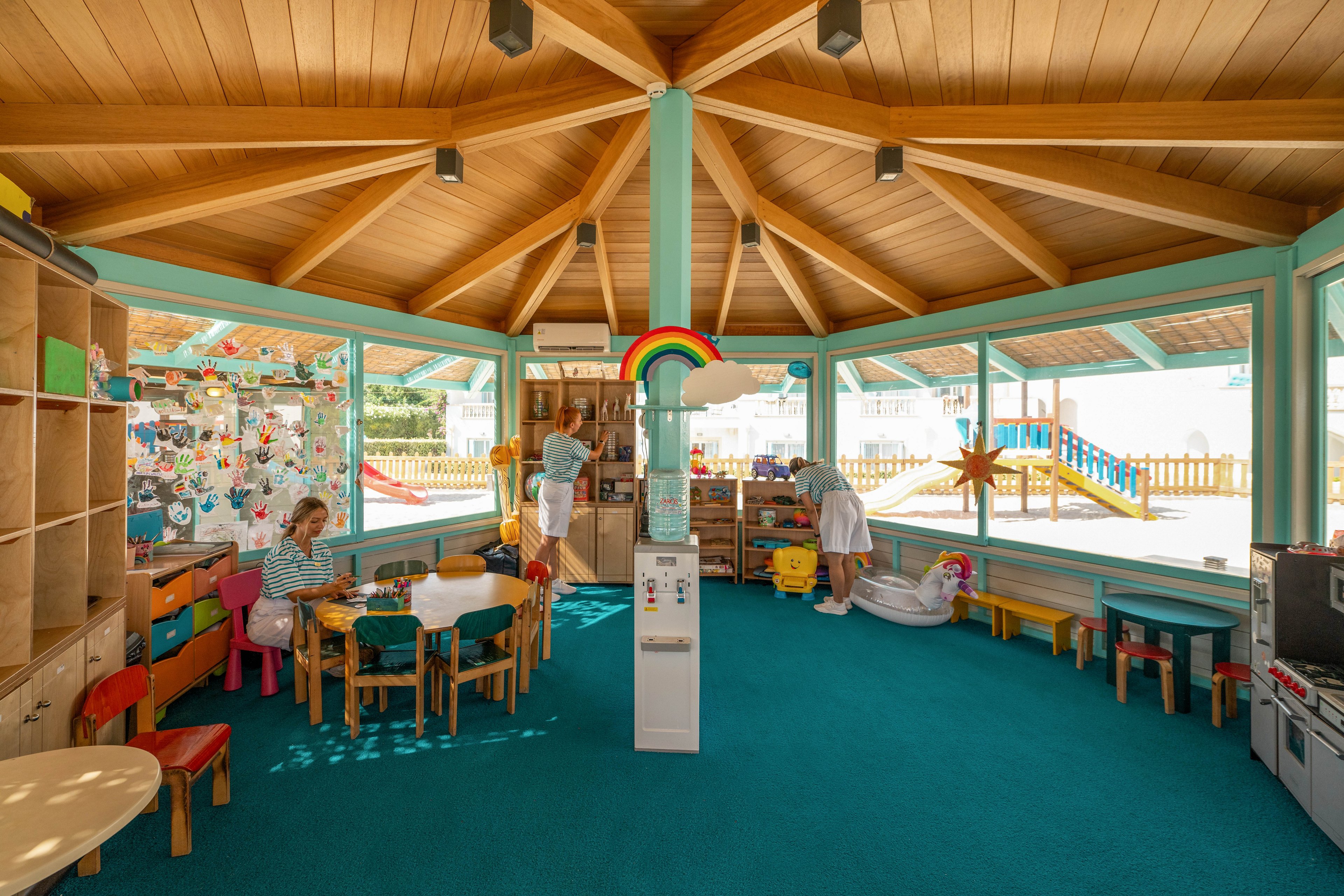 A spacious, sunlit playroom features children engaged in various activities around small tables and play areas. The room has a wooden ceiling with a central column and large windows that let in plenty of natural light. Colorful chairs, shelves filled with toys, and wall decorations create a cheerful, child-friendly environment. A rainbow decoration and outdoor playground visible through the windows enhance the playful atmosphere.