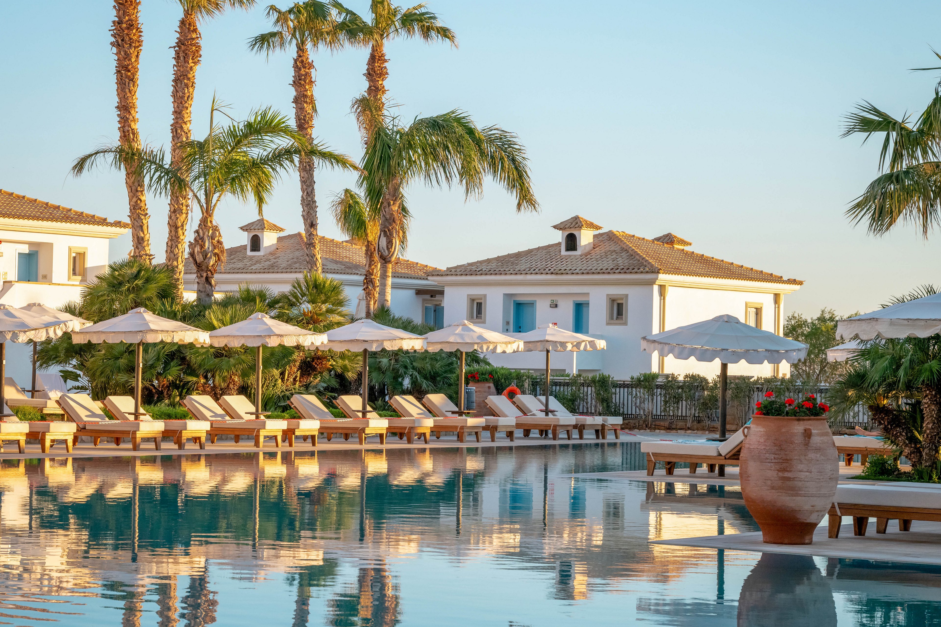A tranquil resort swimming pool reflects rows of white sun loungers and umbrellas in the soft evening light. Mediterranean-style villas with white walls and blue accents line the background, framed by tall palm trees. The calm water surface mirrors the architecture and sky, enhancing the serene atmosphere. A large terracotta pot with flowers adds a decorative touch near the poolside, emphasizing the relaxed vacation setting.