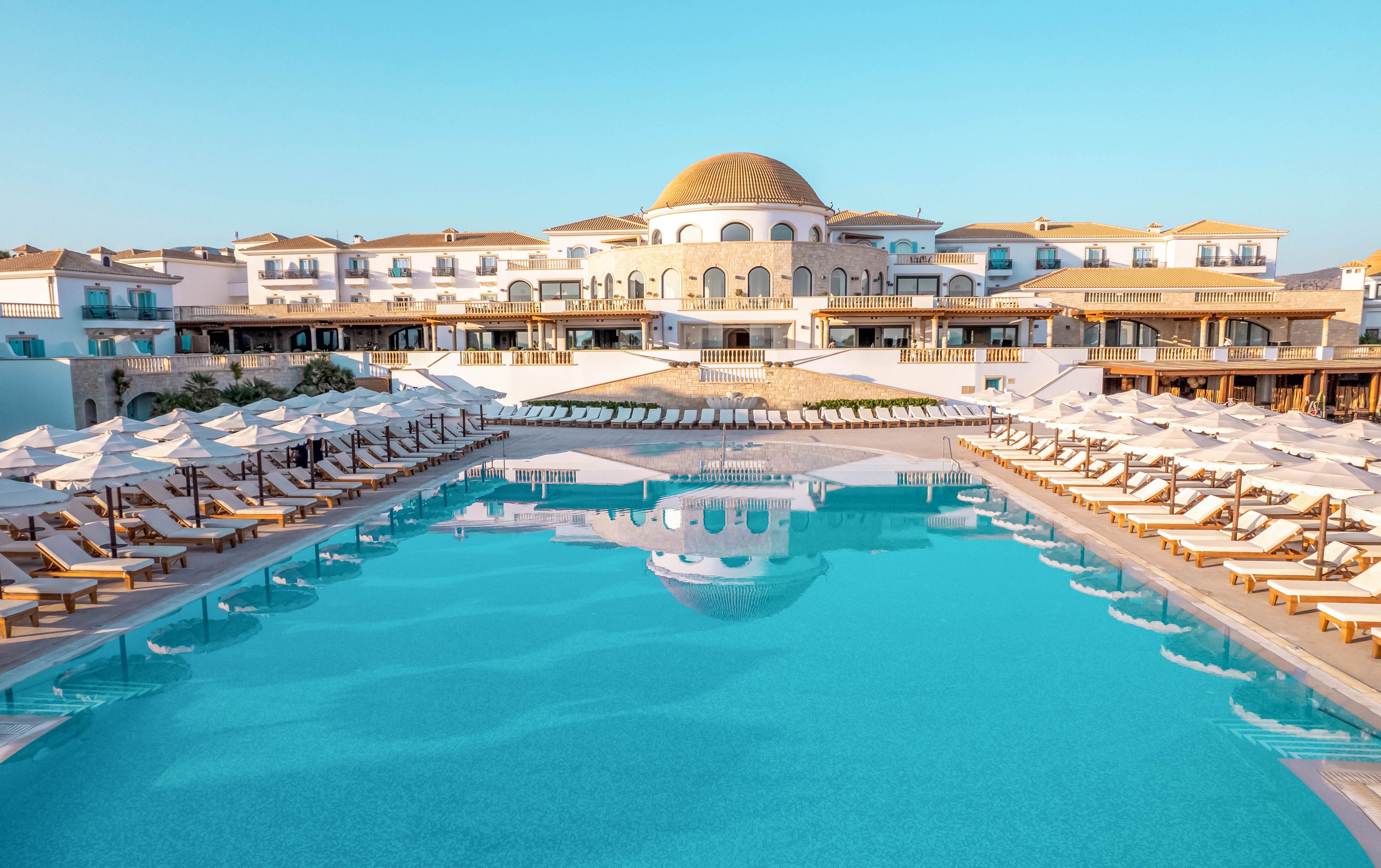 A large turquoise swimming pool dominates the foreground of a luxury resort. Rows of white sun loungers and umbrellas line both sides of the pool, creating a symmetrical and orderly scene. In the background, a grand hotel building with a central dome and multiple balconies stands under a clear blue sky. The warm morning or late afternoon light enhances the tranquil and inviting atmosphere of the resort.