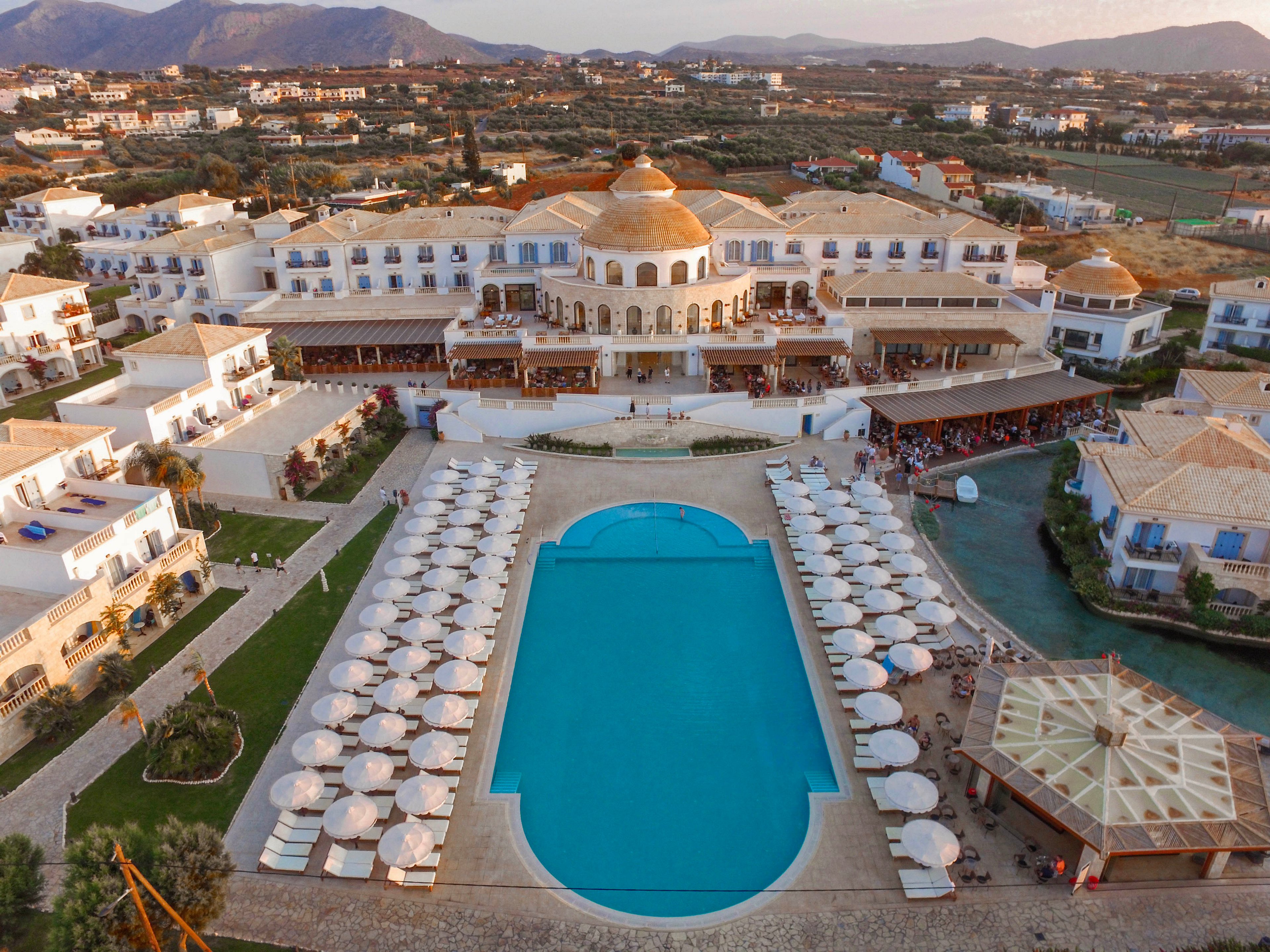 An expansive resort complex is captured from above, featuring a large central swimming pool surrounded by rows of sun loungers and umbrellas. The main building has multiple levels with arched windows and a prominent domed structure at its center. Smaller guest buildings flank the pool area, creating a symmetrical layout. In the background, a rural landscape with scattered houses and distant mountains frames the scene, suggesting a tranquil vacation destination.