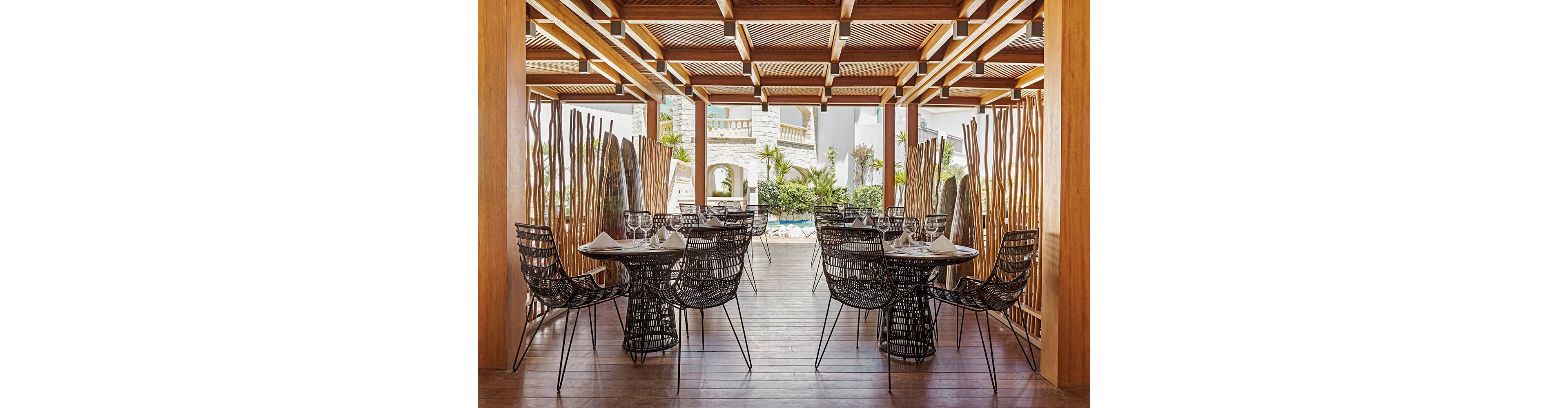 An elegant outdoor dining area features several black wire chairs and round tables set with glassware and folded napkins. The space is framed by warm wooden beams and slatted ceiling panels that create a structured, contemporary look. Decorative vertical wooden sticks and tall plants separate the terrace from the lush garden and pool area beyond. Bright natural light and neutral tones give the scene a relaxed, upscale resort atmosphere.