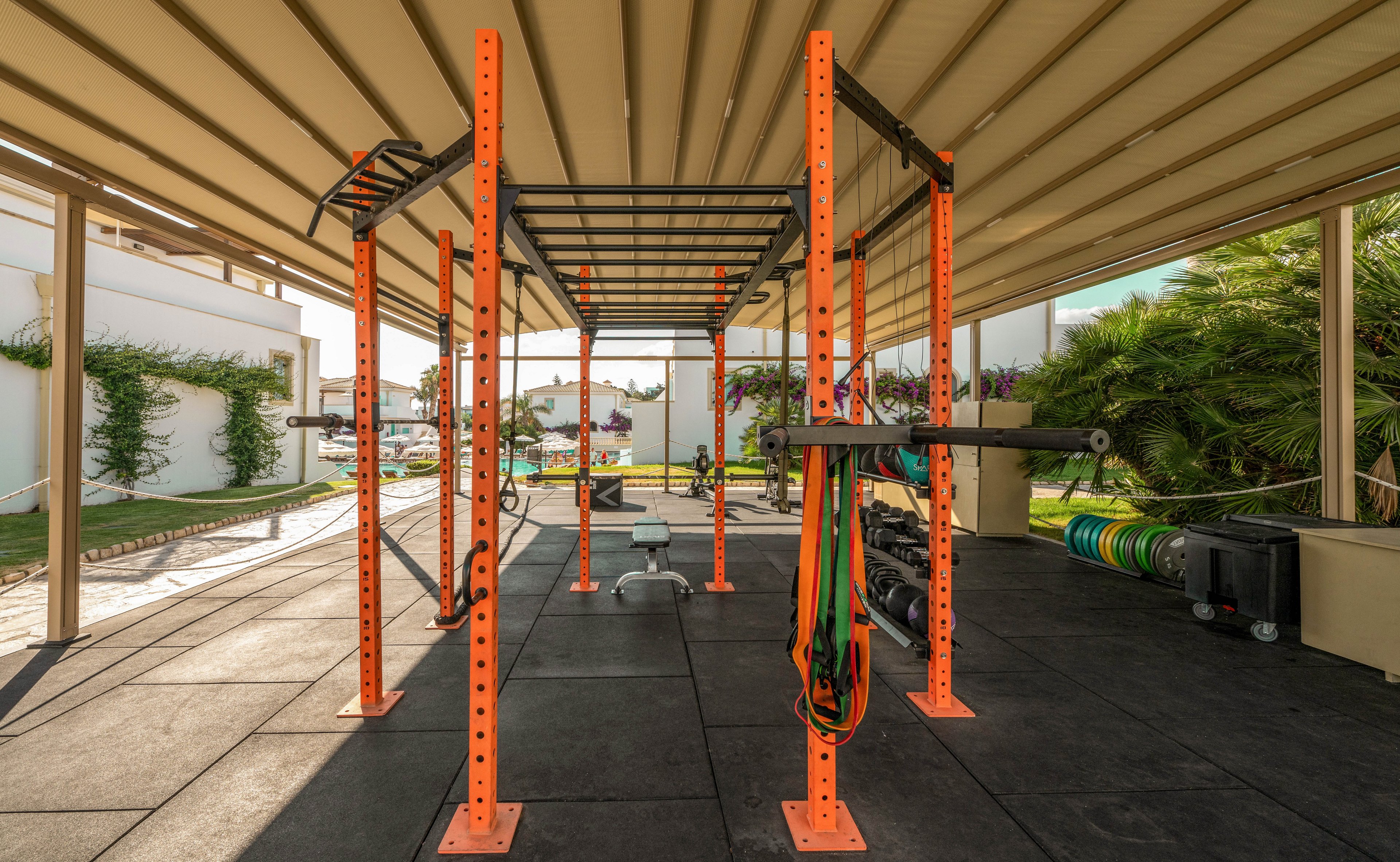 An orange metal fitness rig stands in the center of an outdoor gym area. The structure is placed under a covered canopy with rubber flooring and open sides. Resistance bands and other workout accessories hang from the frame, ready for use. In the background, greenery, palm trees, and residential-style buildings create a bright, sunny setting.