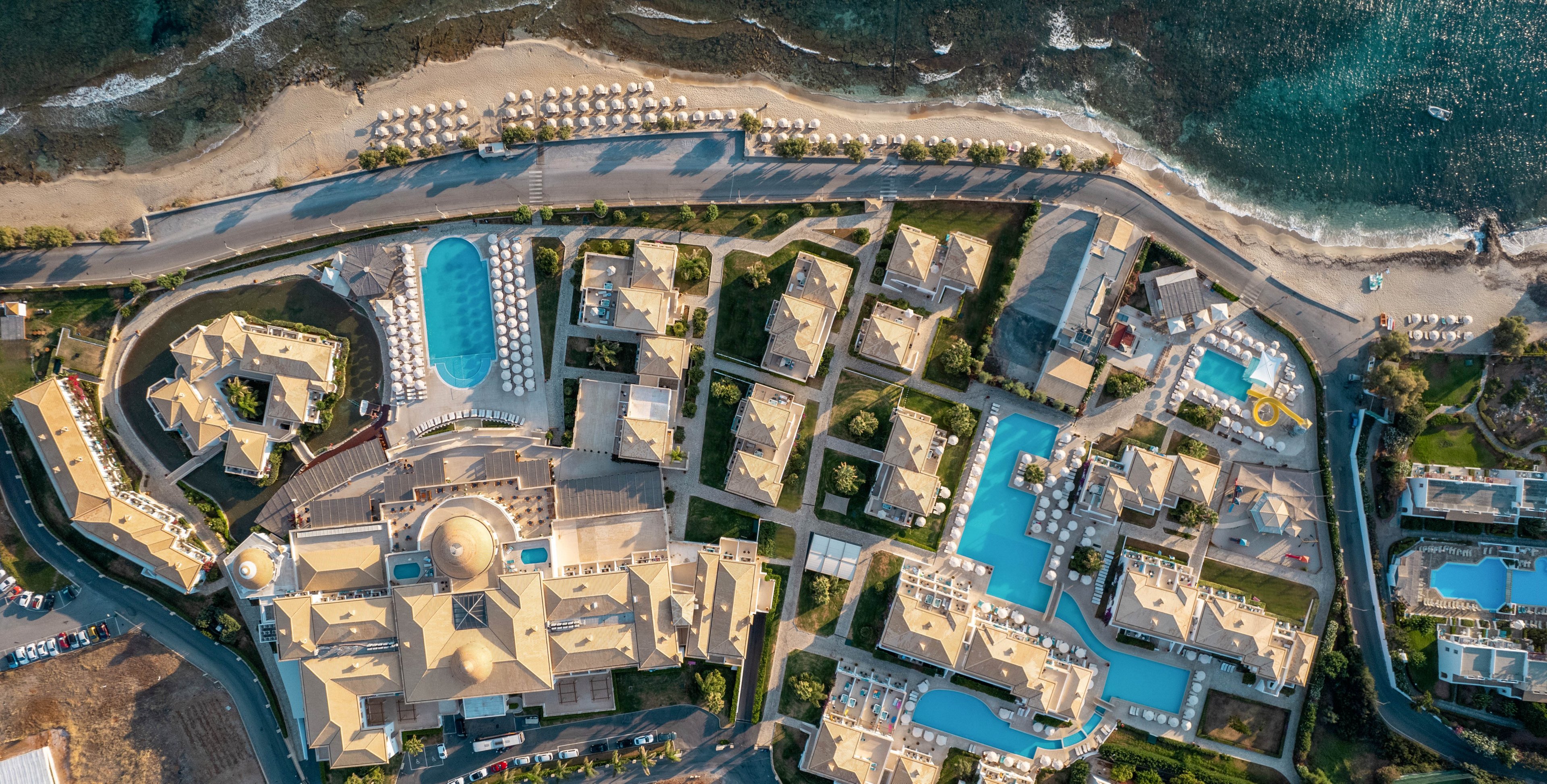 A large seaside resort complex is captured from above, revealing multiple buildings arranged around several bright blue swimming pools. The property stretches along a rocky shoreline with a sandy beach and rows of sun loungers near the water. Curving roads and pathways connect the villas, main hotel structure, and recreational areas. The overall scene suggests a sunny, tranquil vacation destination on the coast.