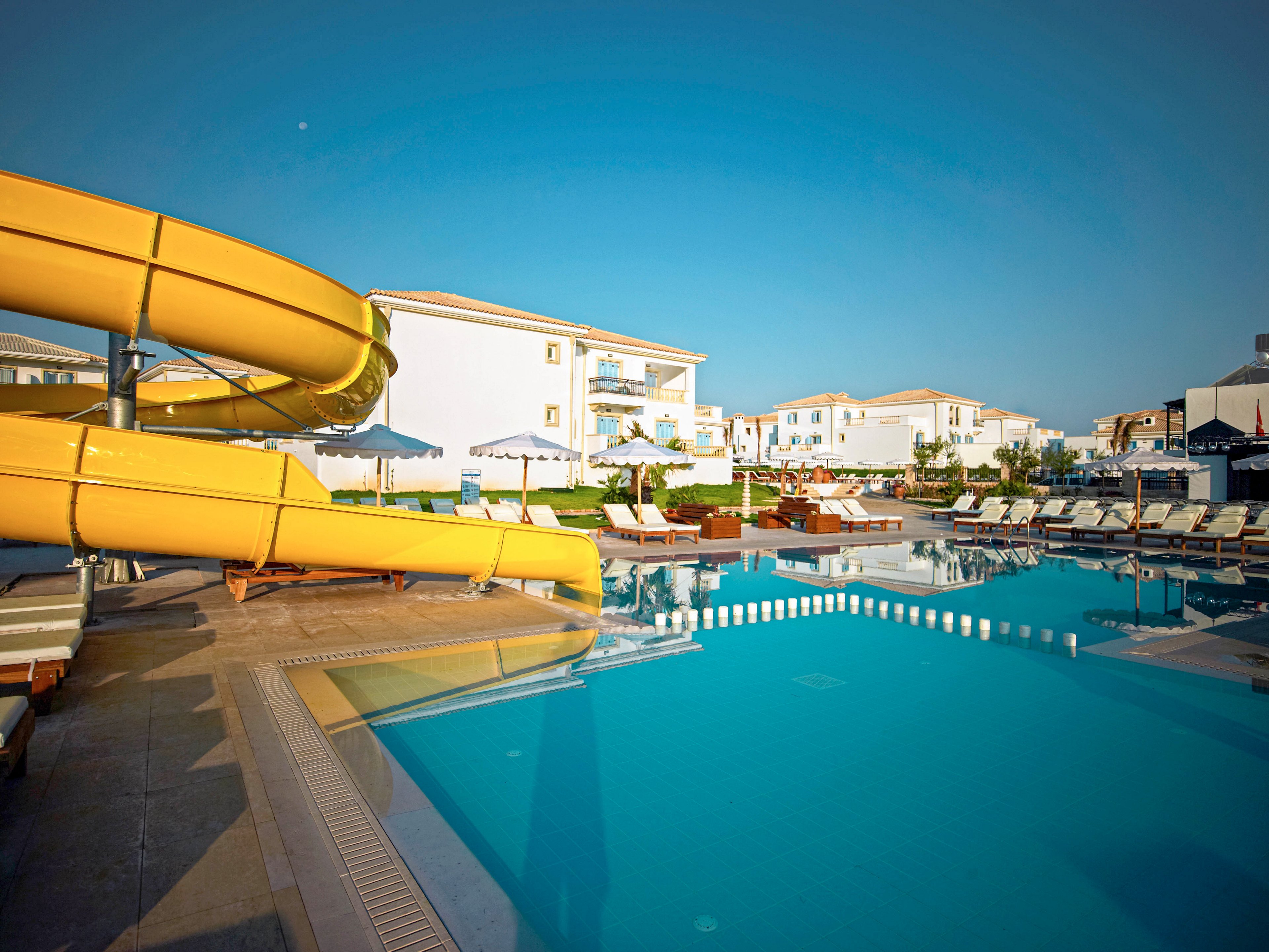 A large outdoor swimming pool dominates the foreground of a modern resort. A bright yellow curved waterslide descends toward the pool, adding a playful feature to the scene. Sun loungers and umbrellas line the poolside, suggesting a relaxing vacation atmosphere. White low-rise buildings with balconies stand in the background under a clear blue sky.
