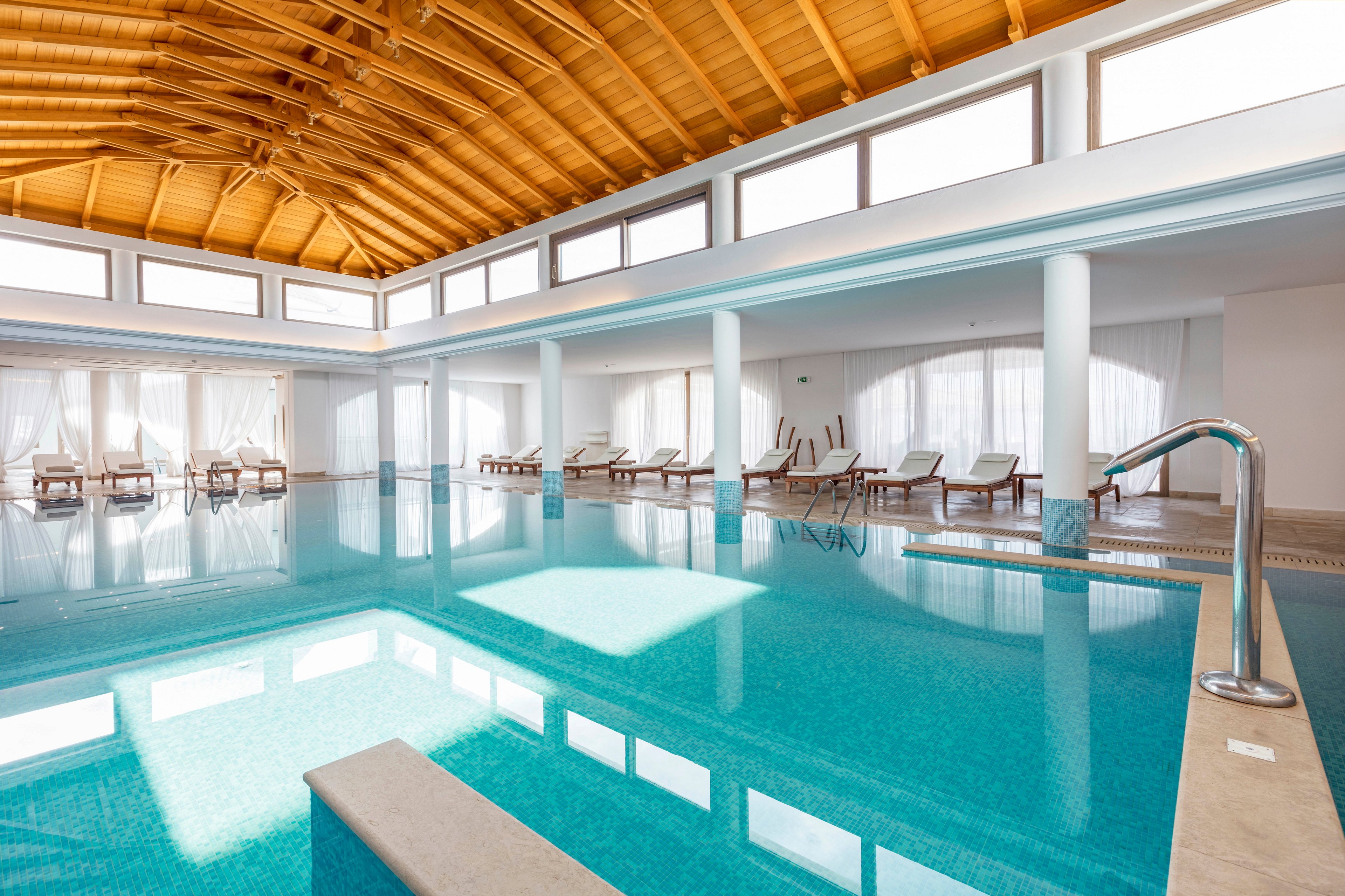 A spacious indoor swimming pool is featured in a modern spa or resort setting. The pool is surrounded by white columns, large windows, and sheer curtains that let in abundant natural light. Wooden ceiling beams add warmth to the bright, minimalist architecture. Several empty lounge chairs line the perimeter, creating a calm and relaxing atmosphere.