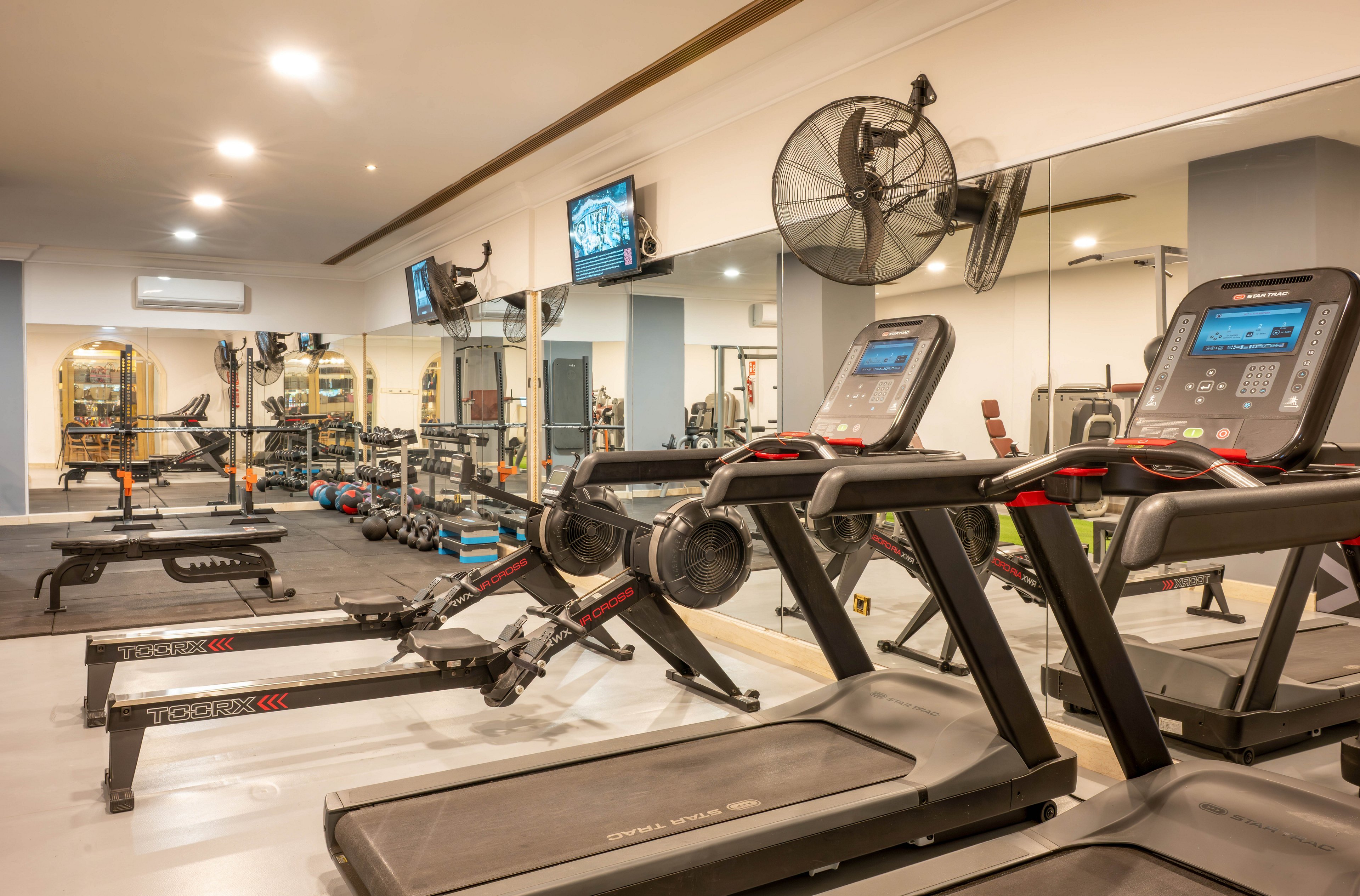 A modern indoor gym features several treadmills in the foreground facing a mirrored wall. In the background, weight machines, benches, and free weights are neatly arranged on rubber flooring. Wall-mounted televisions and a large fan provide entertainment and ventilation for users. Bright, warm lighting and neutral colors create a clean, inviting fitness environment.