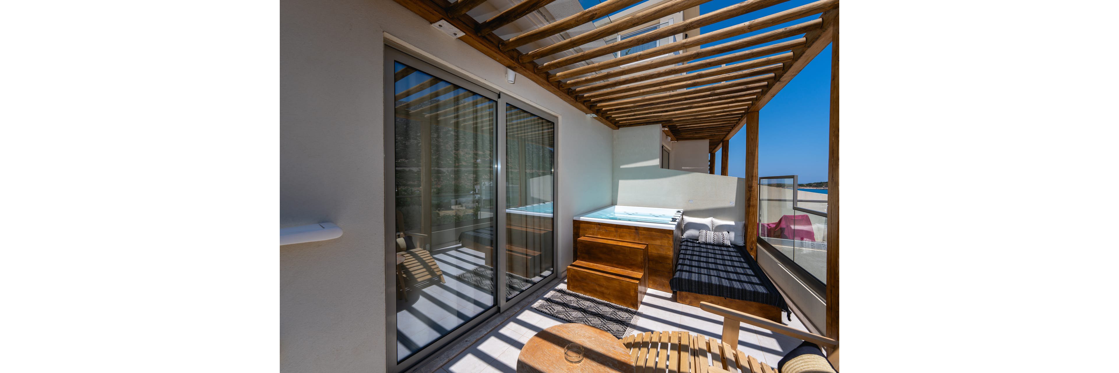 A contemporary outdoor terrace features a wooden hot tub and a cushioned sunbed beneath a slatted pergola. Large sliding glass doors connect the terrace to the interior space, reflecting the bright blue sky. A small wooden table and chair provide a spot for relaxation, while minimalist white walls enhance the clean, modern aesthetic. The scene suggests a private, tranquil retreat with views over the surrounding area.