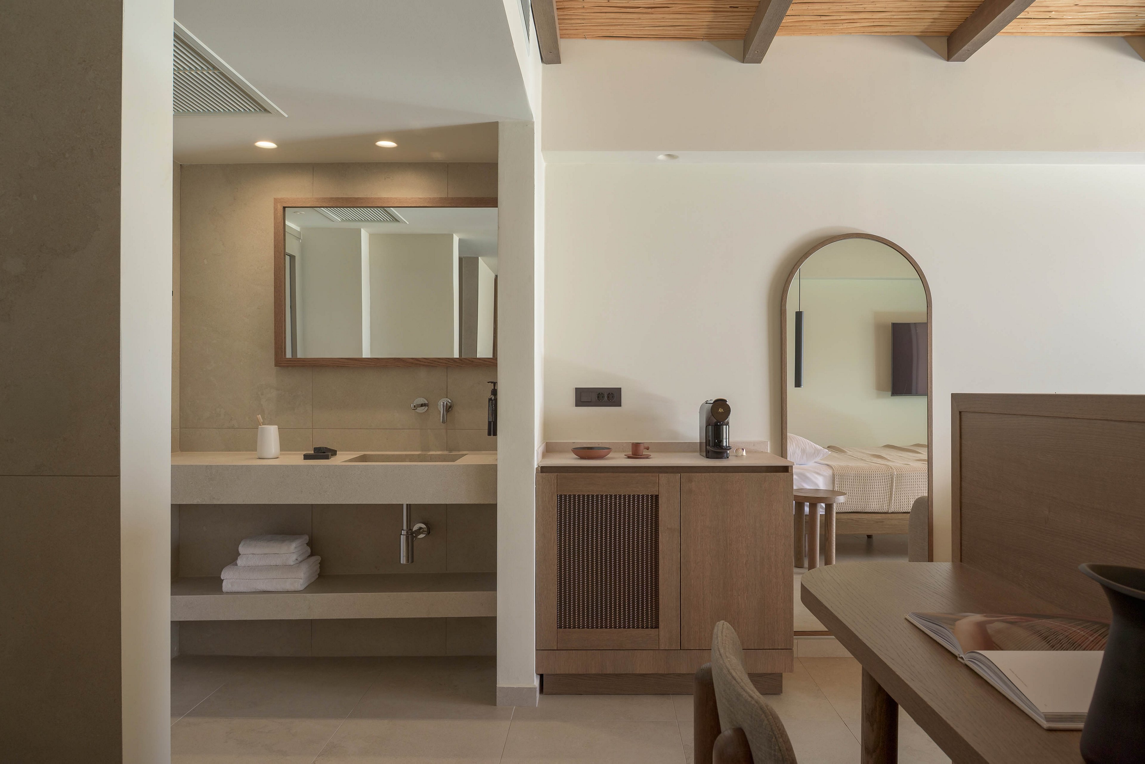 A contemporary hotel suite features a minimalist bathroom with a wide countertop, built-in sink, and neatly folded white towels on an open shelf below. Warm wood cabinetry and a small coffee station sit beside the bathroom area, visually connecting to the adjoining bedroom. The bedroom is partially visible through an arched mirror and doorway, showing a neatly made bed and soft neutral textiles. Recessed ceiling lights and natural daylight create a calm, inviting atmosphere with beige and wood tones throughout.
