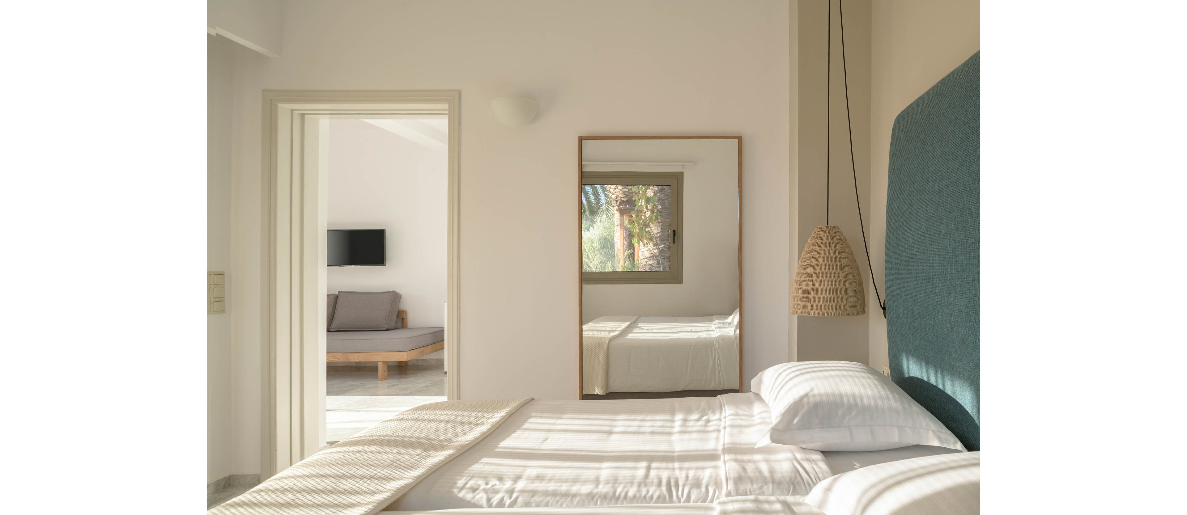 A bright minimalist bedroom features a neatly made bed with white linens and a teal upholstered headboard. Natural light streams in through a window reflected in a large wall mirror, revealing greenery outside. A woven pendant lamp hangs beside the bed, adding a touch of texture to the clean, modern space. Through an open doorway, a sitting area with a sofa and wall-mounted television is visible, continuing the calm, neutral aesthetic.