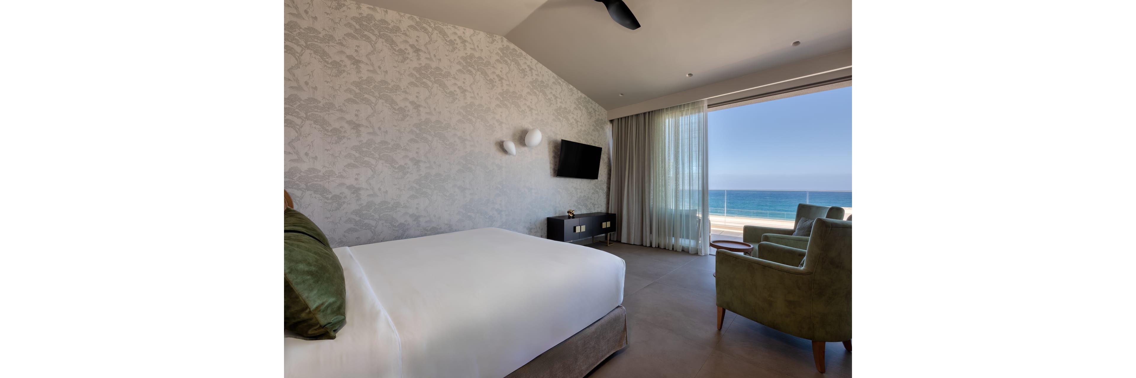 A contemporary hotel bedroom features a large bed with white linens and a green accent pillow. The room includes a wall-mounted television, minimalist nightstand, and textured wallpaper. Floor-to-ceiling curtains open to a balcony overlooking a clear blue ocean horizon. Two upholstered armchairs and a small round table create a cozy seating area facing the view.