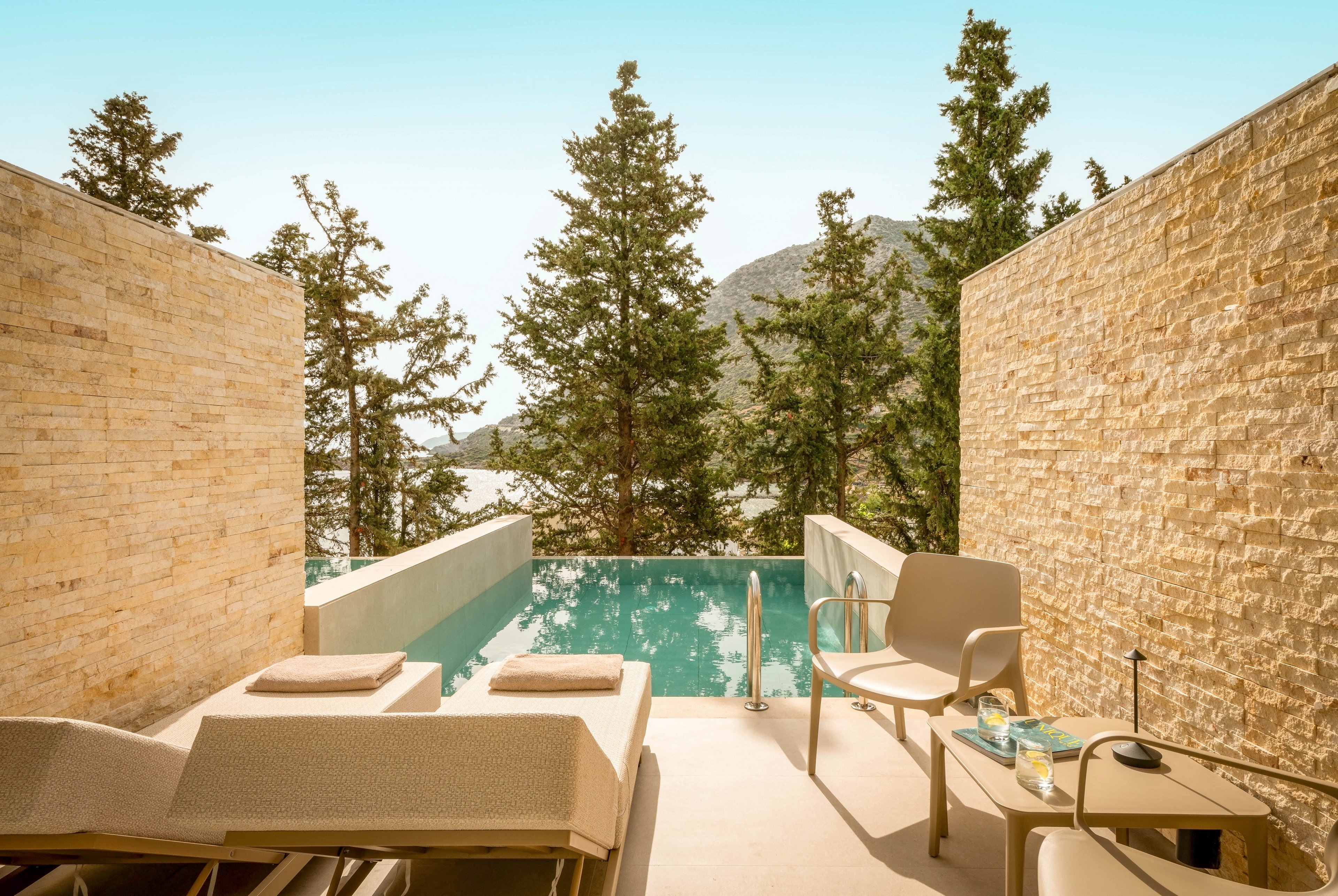 A modern private terrace features two cushioned loungers facing a small turquoise plunge pool. Minimalist outdoor chairs and a side table with drinks create a relaxed seating area. Tall stone walls frame the view of evergreen trees and distant hills under a clear sky. The warm sunlight and neutral tones evoke a serene, upscale resort atmosphere.