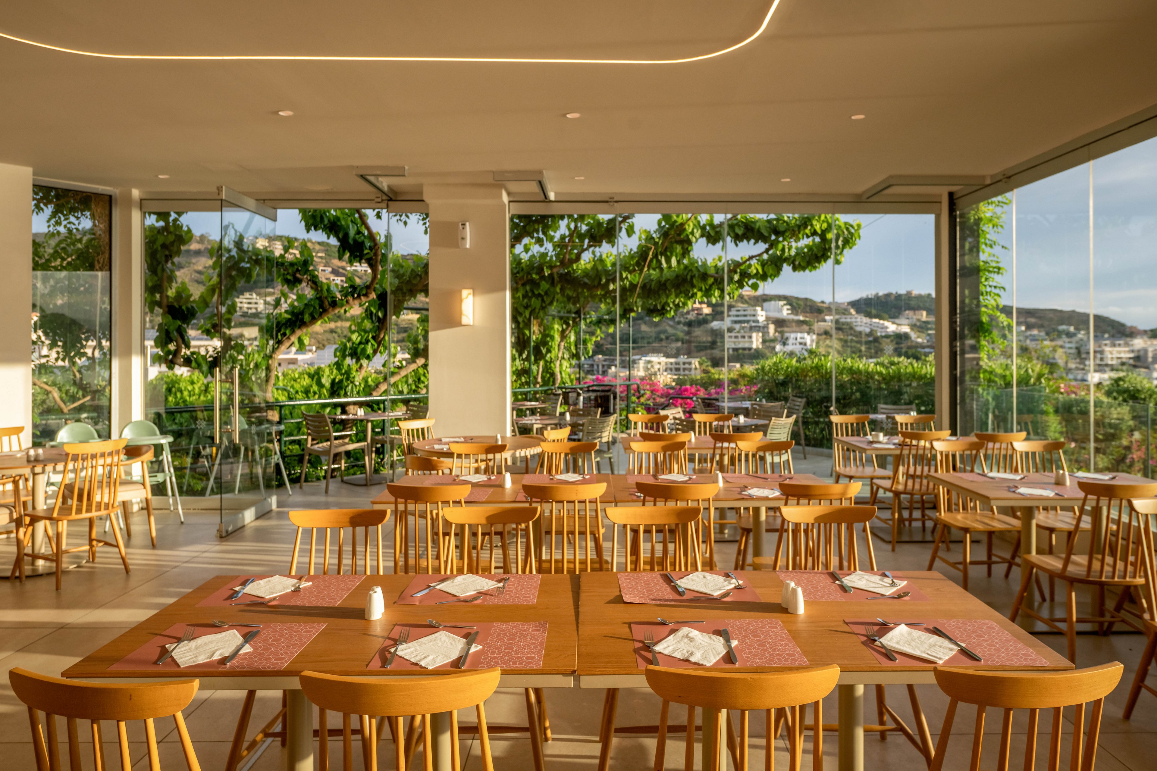 A bright, modern restaurant interior features neatly arranged wooden tables and chairs. Place settings with napkins and menus are prepared on each table, suggesting readiness for guests. Large floor-to-ceiling glass windows open to lush greenery, flowering plants, and a distant hillside town. Warm natural light fills the space, creating an inviting and relaxed dining atmosphere.