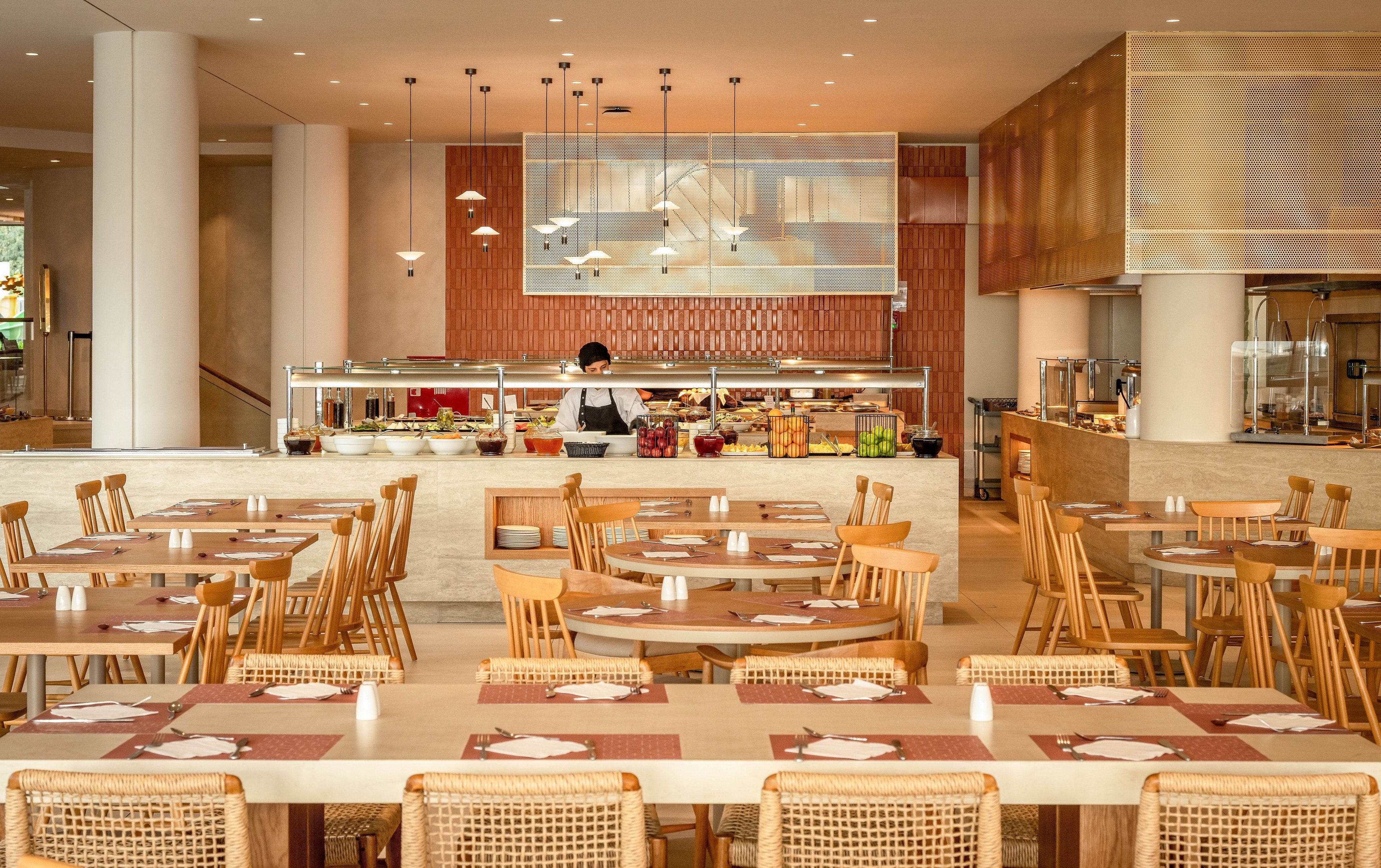 A contemporary restaurant dining area features neatly arranged wooden tables and chairs. In the background, a chef works behind a buffet counter filled with food and drink options. Warm lighting, neutral tones, and clean lines create a welcoming, minimalist atmosphere. The space appears organized and ready for guests, with placemats and table settings prepared on each table.