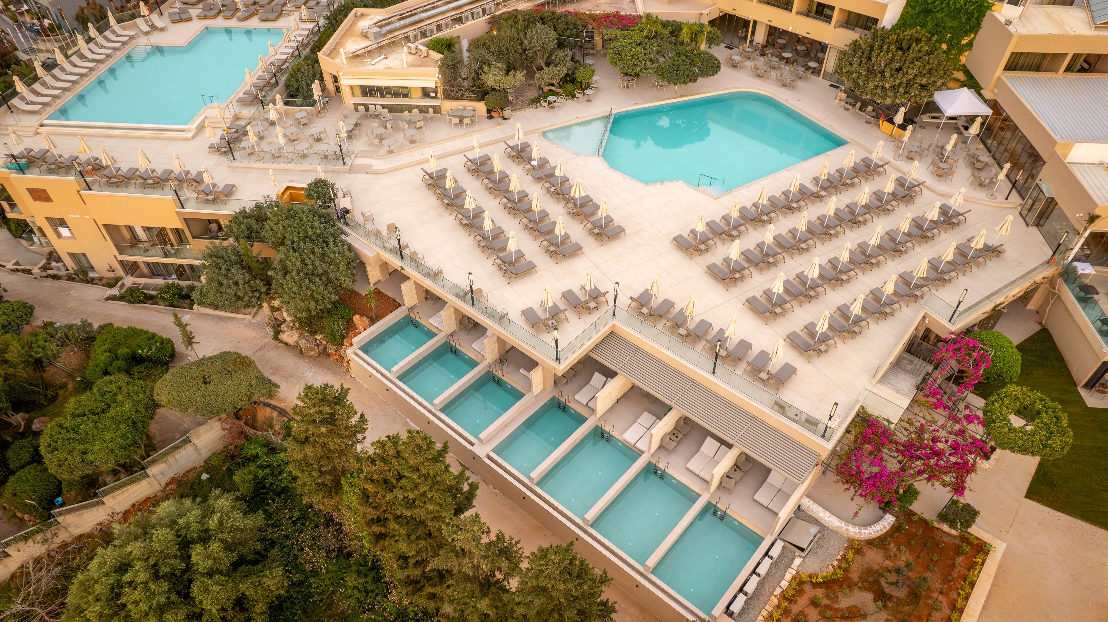 A modern resort complex is captured from above, showcasing multiple swimming pools and sun loungers. The main terrace features rows of neatly arranged lounge chairs around a large pool. Below, several private plunge pools line a lower level with cabana-style seating. Surrounding greenery and flowering plants soften the architecture and create a relaxing vacation atmosphere. The scene suggests a tranquil, upscale holiday destination in a warm climate.