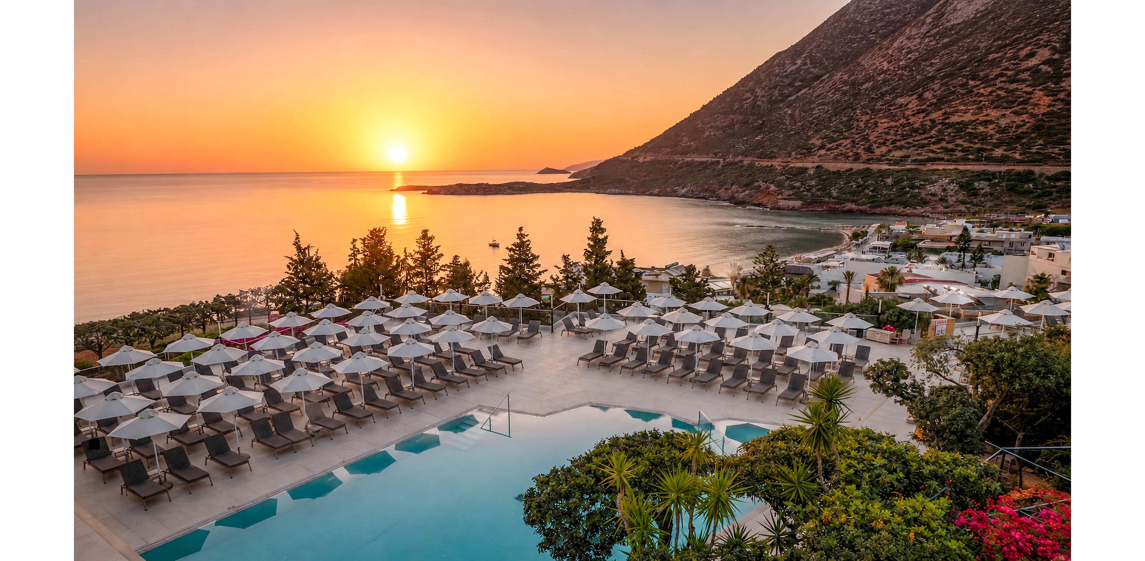 A coastal resort pool is surrounded by neatly arranged sun loungers and umbrellas overlooking a calm sea at sunset. The sun is low on the horizon, casting warm orange and gold tones across the water and sky. A steep mountain rises on the right side, framing the bay and small seaside village below. Lush greenery and flowering plants in the foreground add contrast to the serene, symmetrical pool area.