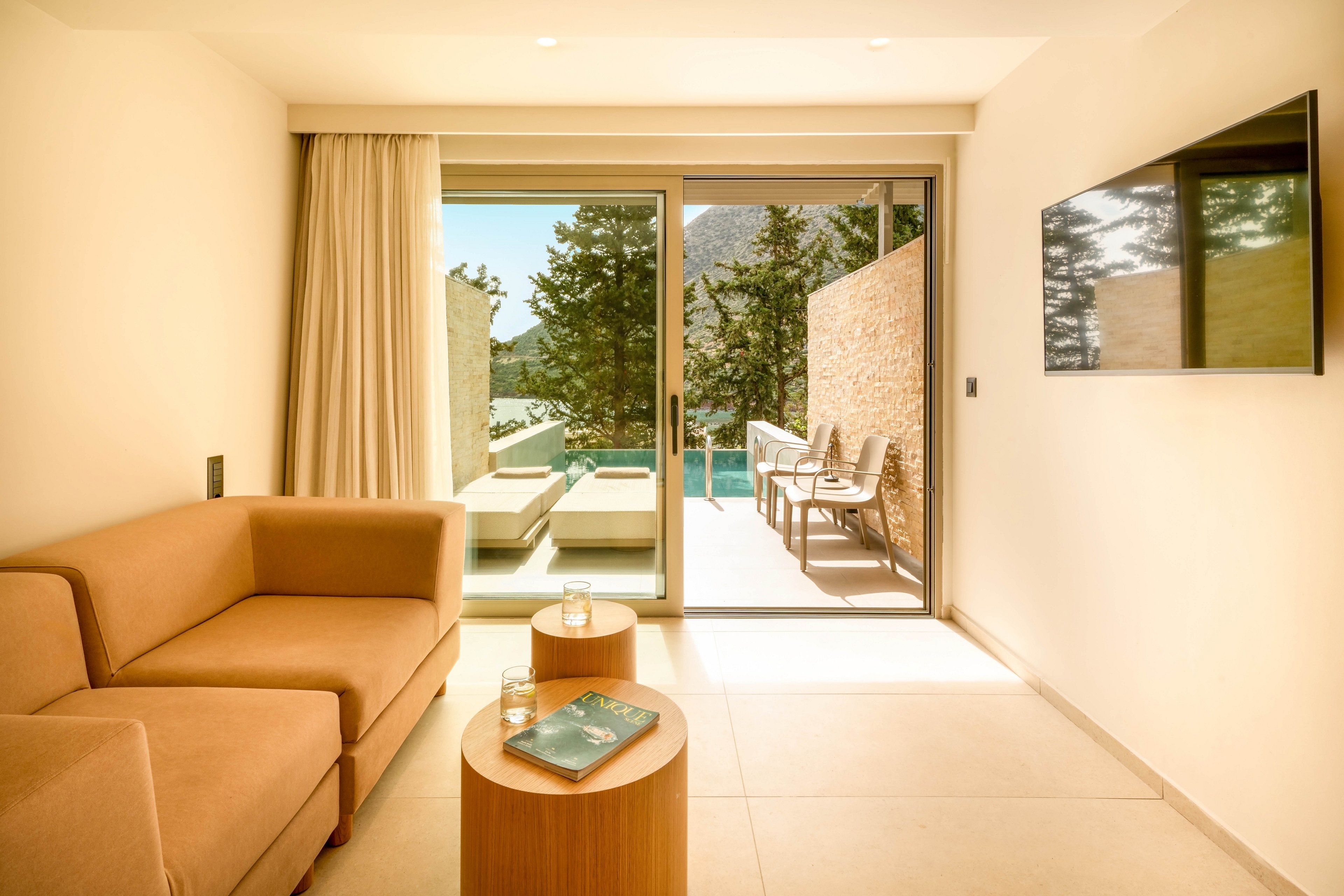 A contemporary hotel room features a tan sofa and round wooden side table in a bright, minimalist interior. Large sliding glass doors open to a private terrace with lounge chairs and a small pool. Sunlight floods the space, highlighting neutral tones and clean lines. A wall-mounted flat-screen TV and outdoor stone wall complete the modern, relaxing atmosphere.