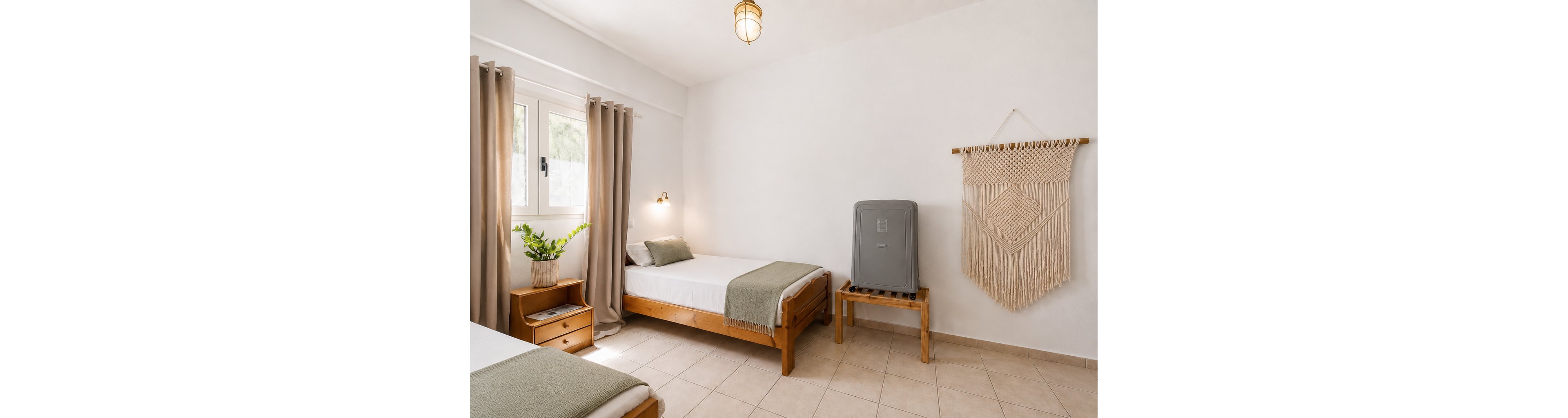A bright minimalist bedroom features two wooden twin beds with white linens and light green throws. Natural light enters through a window framed by beige curtains, illuminating the neutral tiled floor. A potted plant adds greenery on a small bedside table, while a woven wall hanging decorates the white wall. A gray suitcase rests on a wooden stand, suggesting a cozy guest room or vacation rental.
