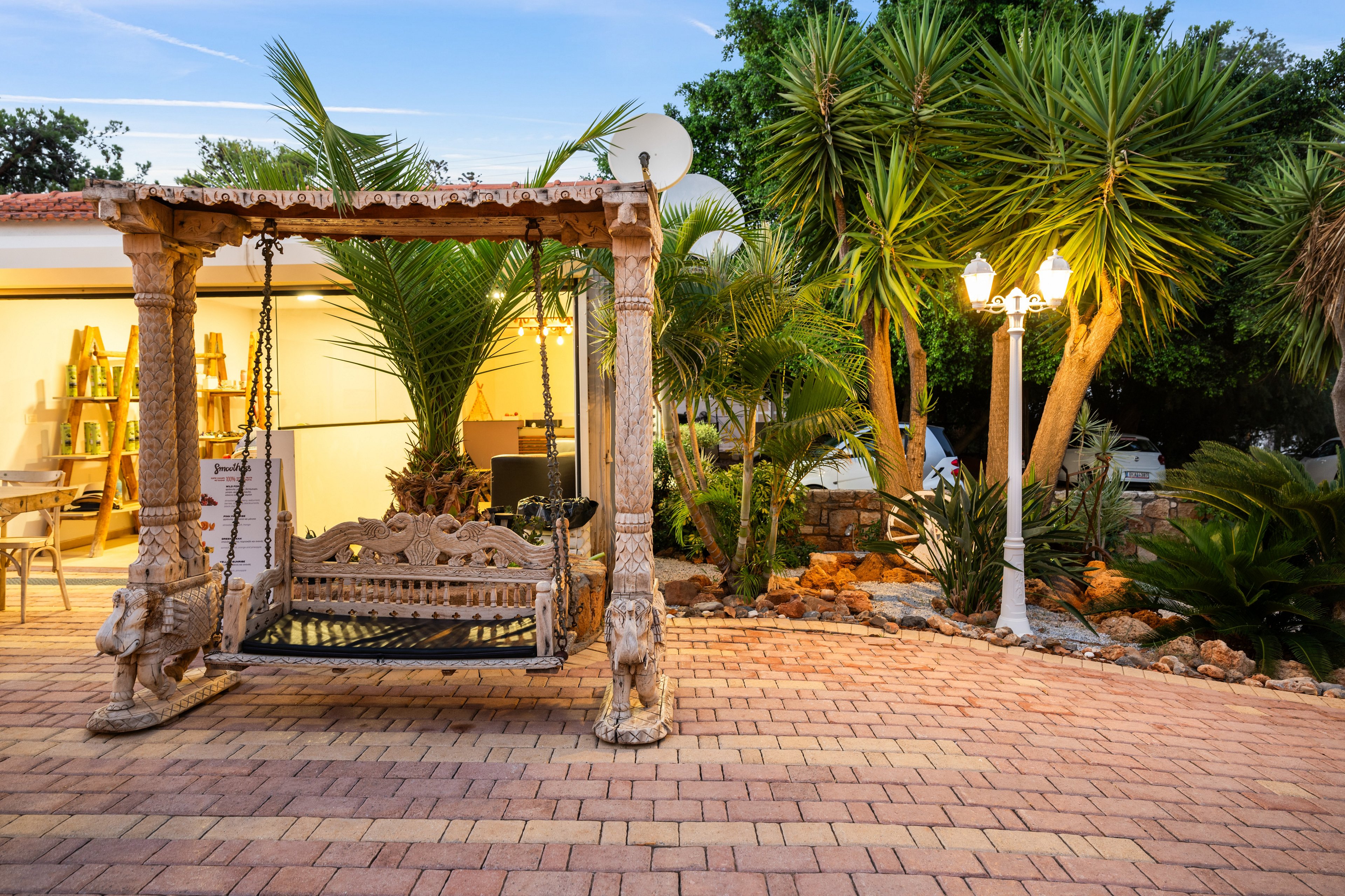An ornate carved wooden swing hangs from a decorative frame on a spacious paved patio. The scene is set at dusk beside a warmly lit house with large open doors. Tropical plants and palm trees surround the seating area, adding lush greenery and texture. A classic white lamppost and stone landscaping elements enhance the cozy, inviting outdoor atmosphere.
