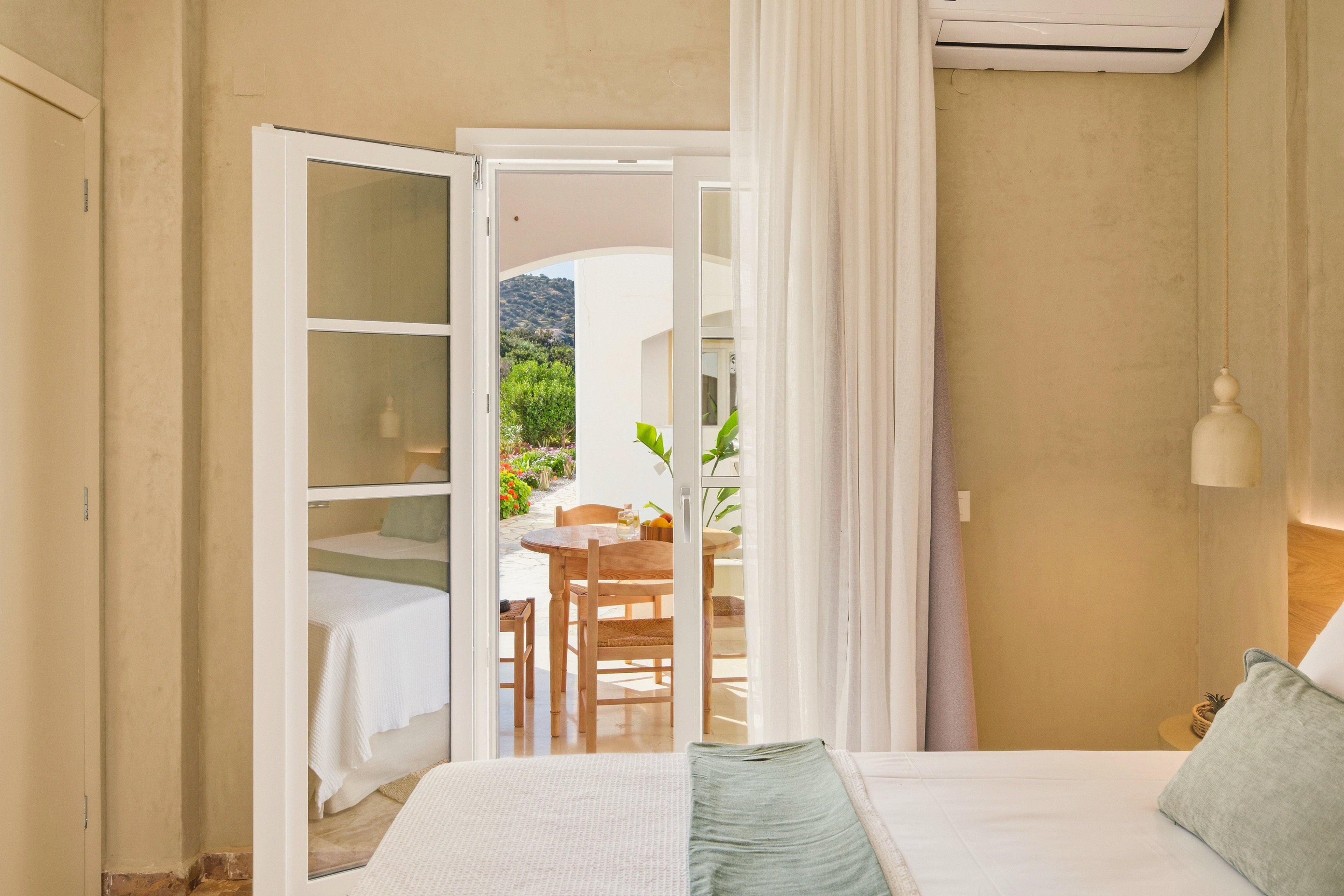 Bright hotel-style bedroom with a neatly made bed opens onto a sunlit terrace through white French doors. Soft neutral walls and light textiles create an airy, minimalist atmosphere. Outside, a wooden table and chairs sit under a covered patio with greenery and distant hills visible. Sheer curtains and a wall-mounted pendant lamp add a relaxed, Mediterranean resort feel.