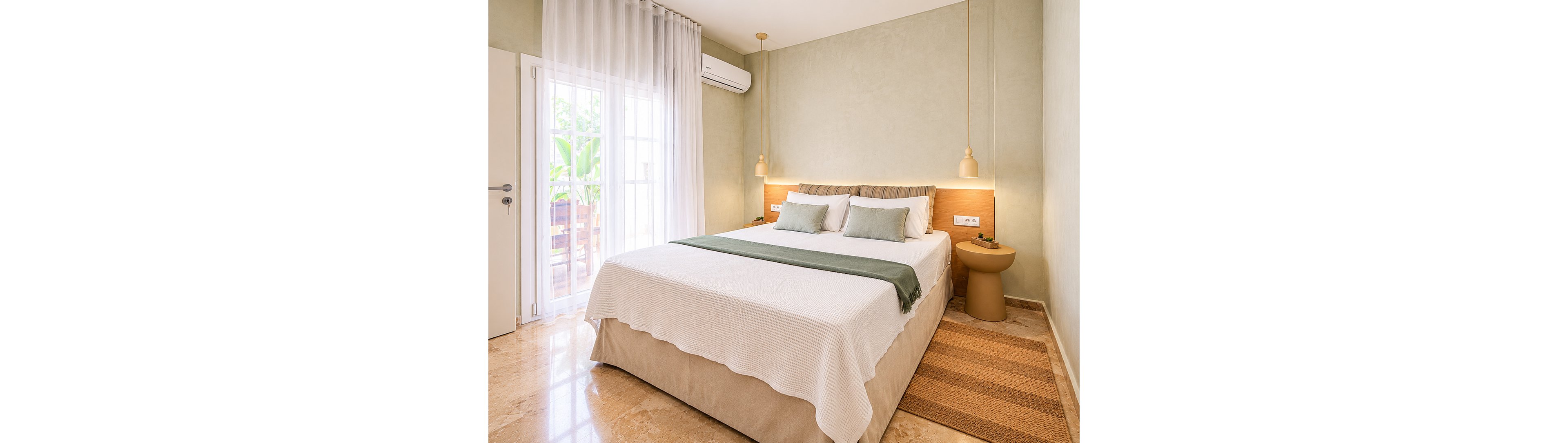 A neatly made double bed with white bedding and a green throw is centered in a bright, minimalist bedroom. Soft beige walls and a matching floor create a warm, neutral palette. Natural light streams in through sheer curtains covering a glass door that opens to an outdoor area. Two pendant lights hang above a built-in headboard shelf, with a small side table and wall-mounted air conditioner completing the modern interior.