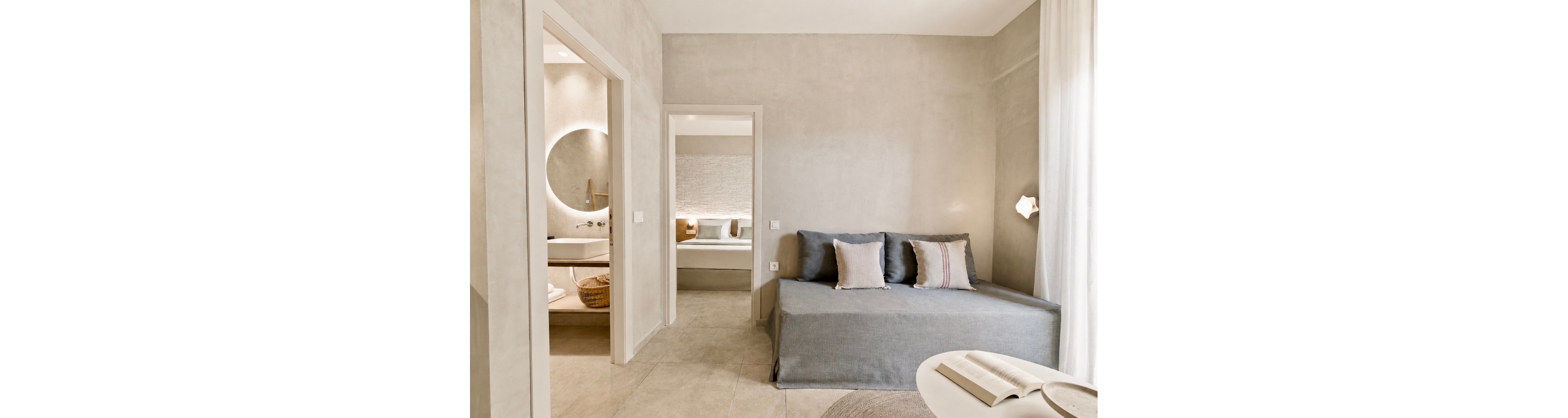 A minimalist hotel-style suite features a gray daybed with neutral accent pillows against smooth plaster walls. In the background, an open doorway reveals a contemporary bathroom with a round illuminated mirror and a woven basket. Soft natural light filters through sheer curtains, enhancing the beige and gray color palette. A small table with an open book and folded textiles adds a relaxed, cozy touch to the modern space.