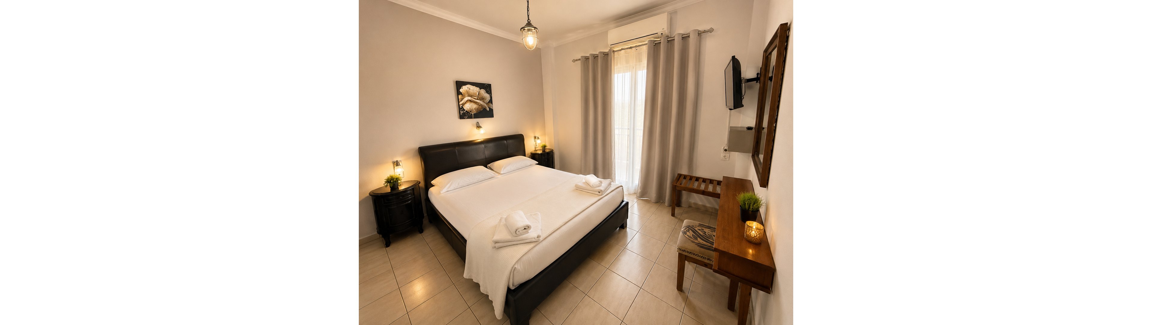A neatly made double bed with white linens is the focal point of this cozy bedroom. The room features warm lighting, neutral walls, and tiled flooring, creating a calm and inviting atmosphere. Sheer curtains cover a large window or balcony door, allowing soft natural light to filter in. Additional details include bedside tables with lamps, a wall-mounted television, a wooden desk with a chair, and a small decorative plant.