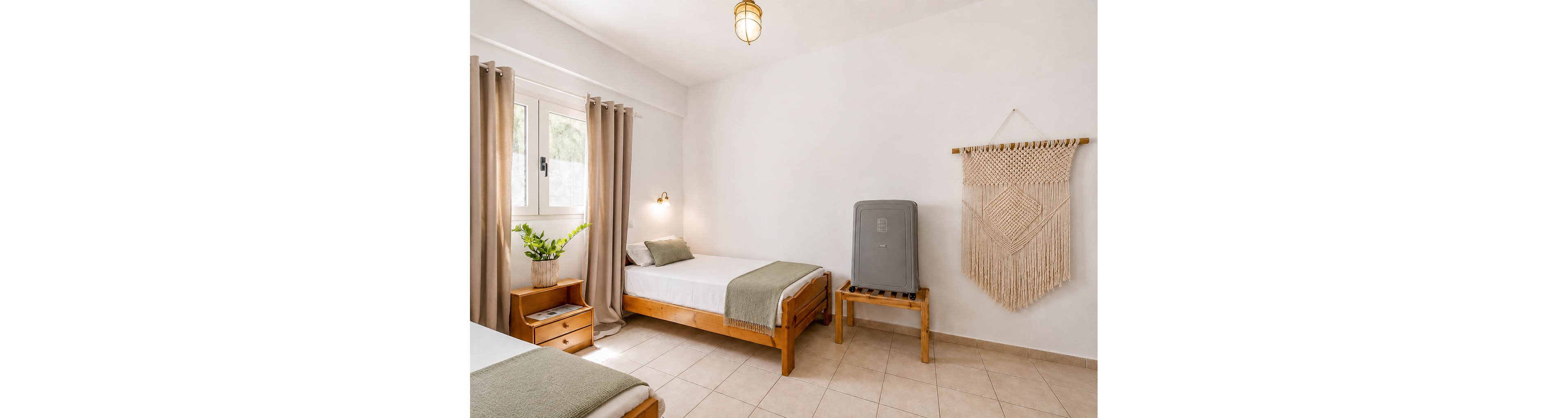 A bright minimalist bedroom features two simple wooden twin beds with white linens and sage green throws. Natural light enters through a window framed by beige curtains, illuminating the neutral tiled floor. A potted plant adds a touch of greenery near the window, while a woven wall hanging decorates the white wall. A small wooden stand holds a gray suitcase or storage case, contributing to the room’s functional, guest-ready feel.
