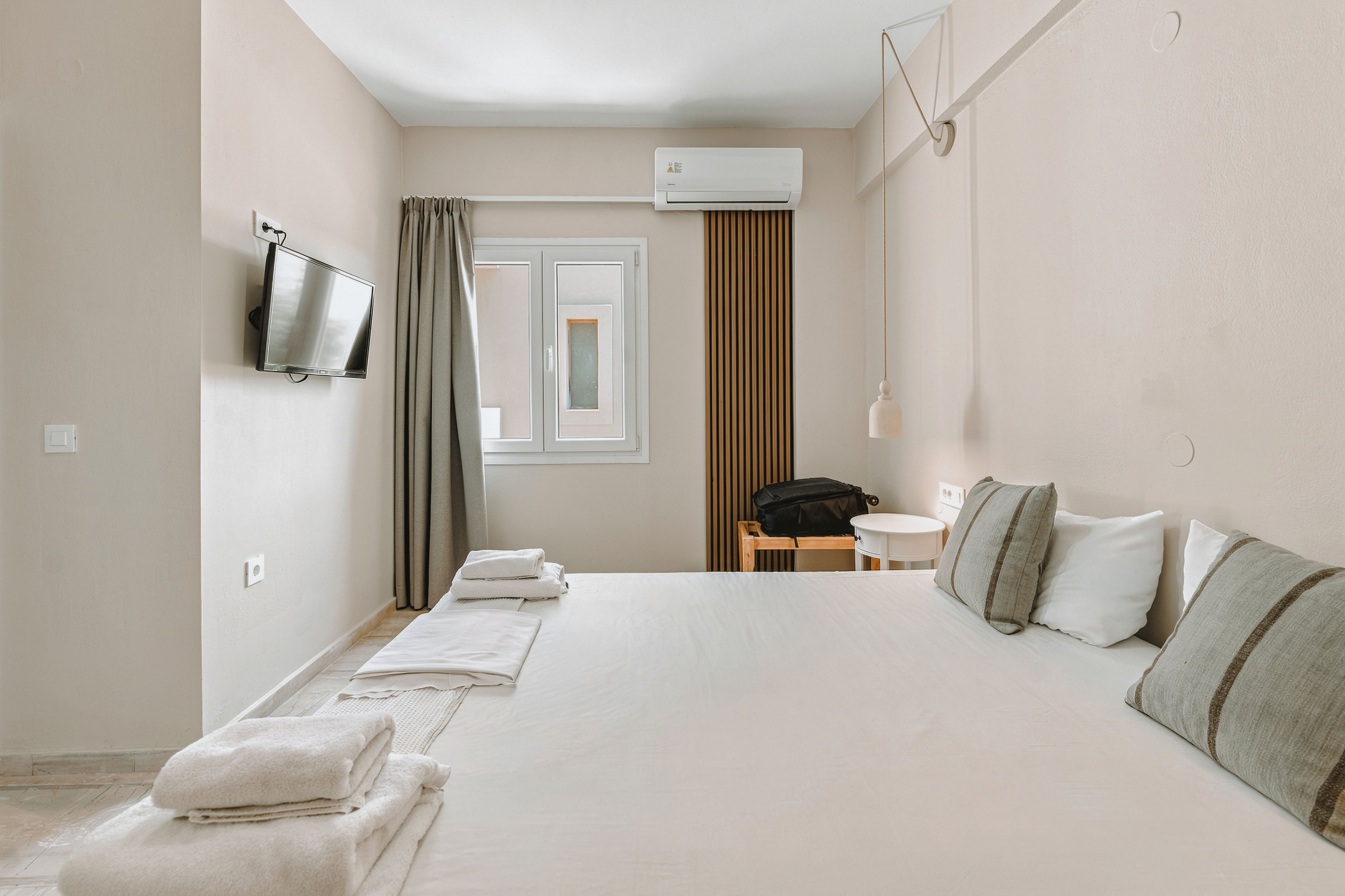 Bright, minimalist bedroom interior featuring a neatly made double bed with white linens and accent pillows. A wall-mounted television and air conditioning unit are visible opposite the bed, along with a window framed by curtains. Soft beige walls and light flooring create a clean, neutral atmosphere. Folded towels on the bed and a small bedside table contribute to a tidy, welcoming hotel room setting.