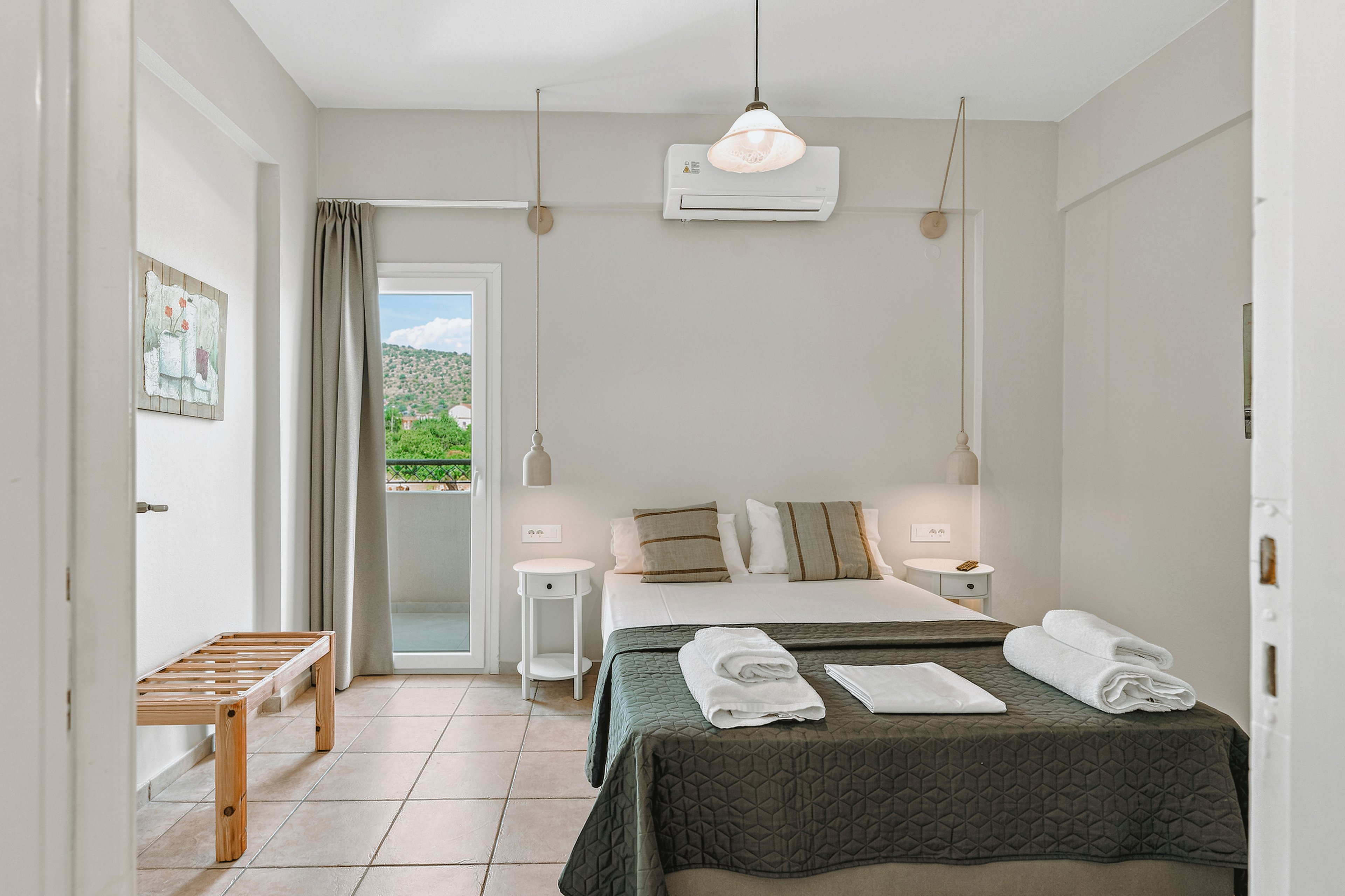 A clean, minimalist hotel bedroom features a double bed neatly arranged with folded white towels and neutral-toned bedding. The room is decorated in soft beige and white colors, with simple pendant lights and a small bedside table on each side of the bed. A wooden bench stands near the doorway, and a glass door opens to a balcony with a scenic view of green hills. The overall atmosphere is calm, airy, and inviting, emphasizing comfort and simplicity.