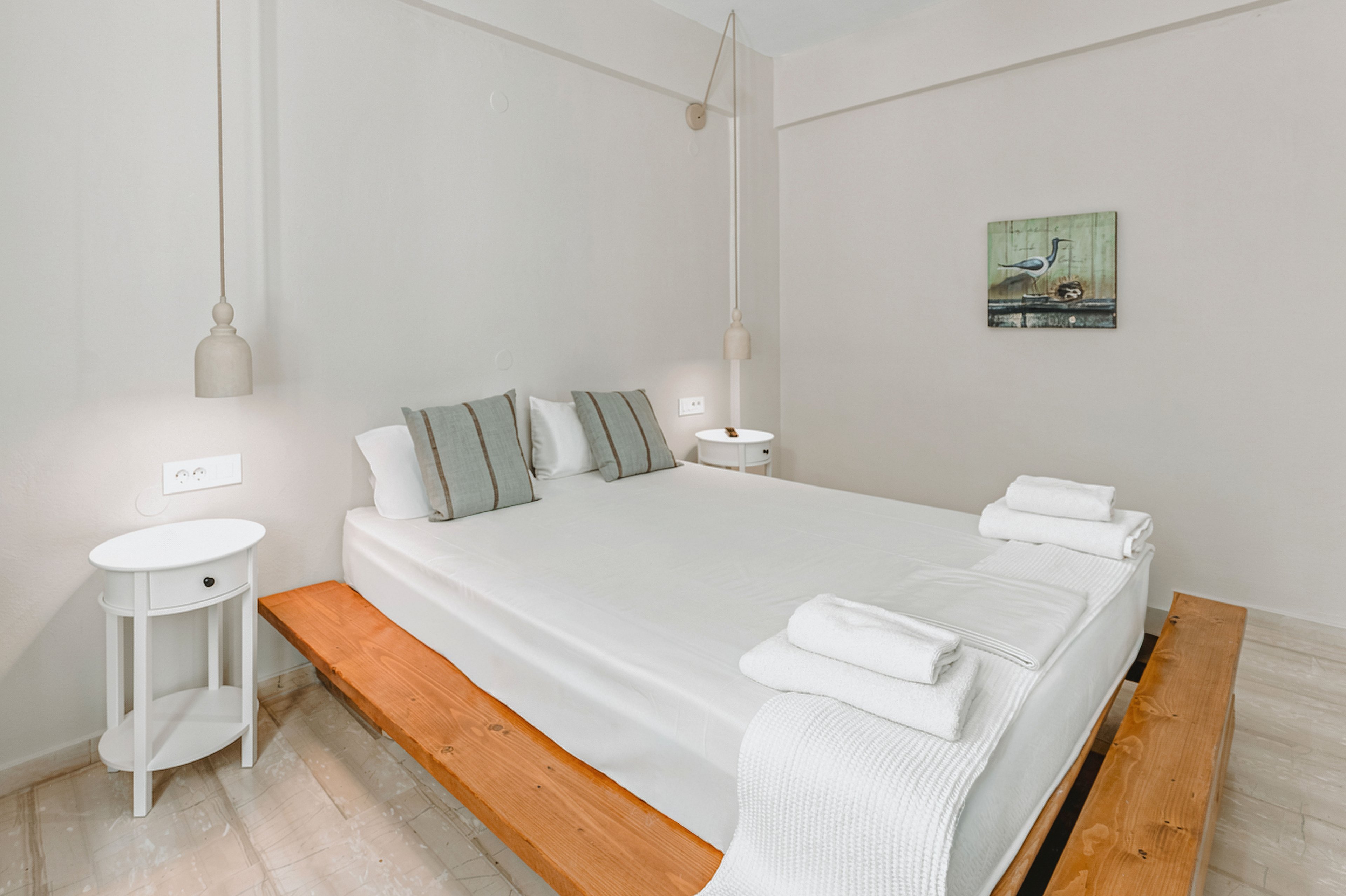 A bright minimalist bedroom features a double bed on a wooden platform with crisp white linens and neatly folded towels. Two small round bedside tables flank the bed, each with a modern hanging pendant light above. Soft neutral walls and flooring create a calm, airy atmosphere. A small framed artwork with a bird scene adds a subtle decorative touch on the wall.